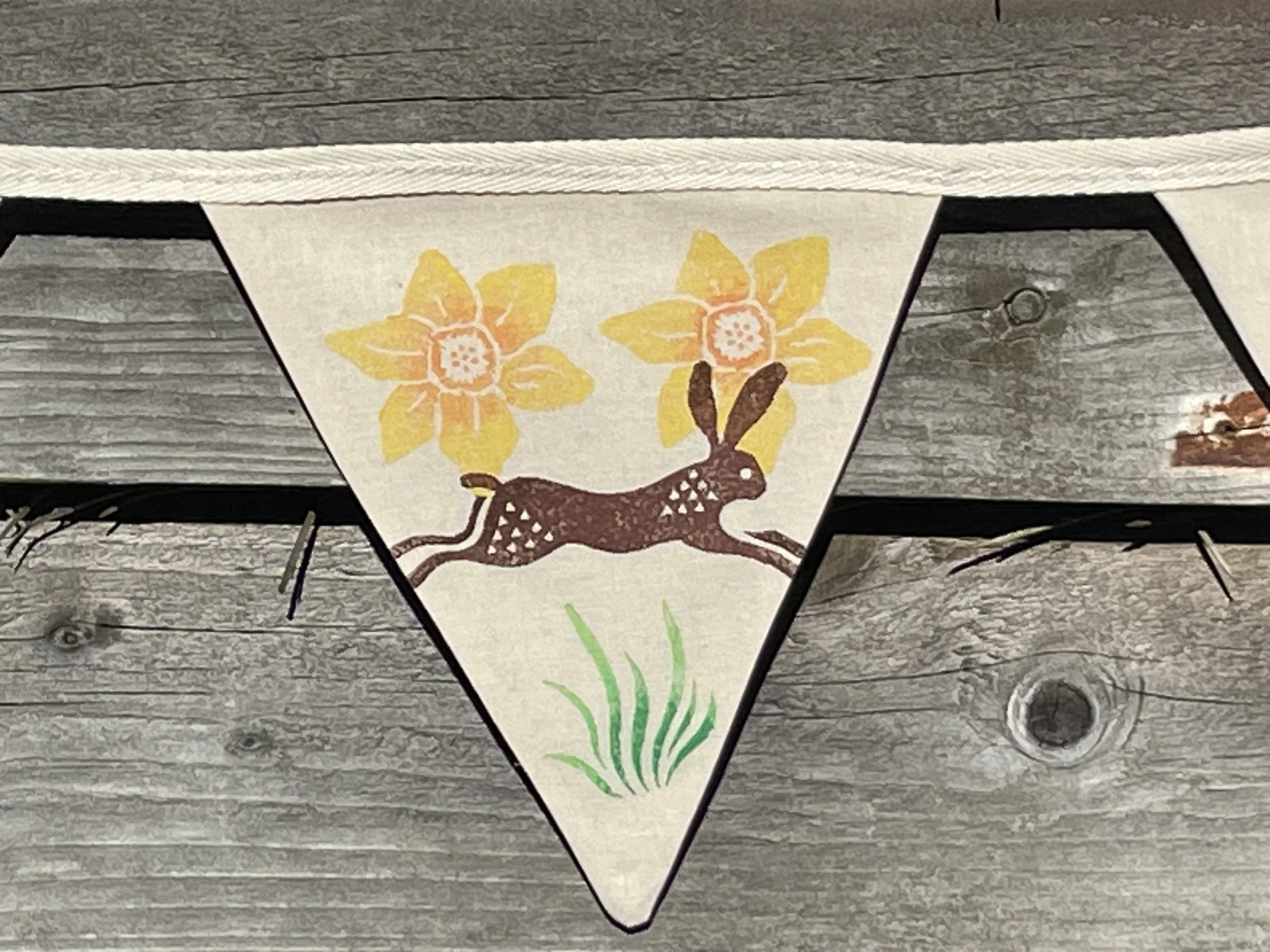 Hand made cotton bunting with woodblock printed yellow flowers, a brown hare and green grass hung on weathered wooden surface.