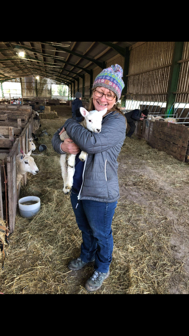 Woman smiling and holding a baby goat inside a barn with other goats in the background.