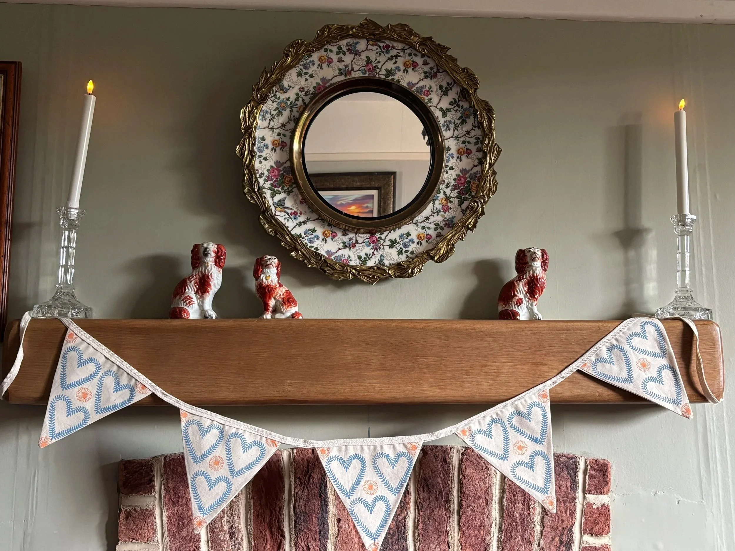 Decorative fireplace mantle with a fabric valance draped across it, three ceramic dog figurines, two tall glass candlesticks with lit candles, and a round ornate floral mirror hanging on the wall behind.