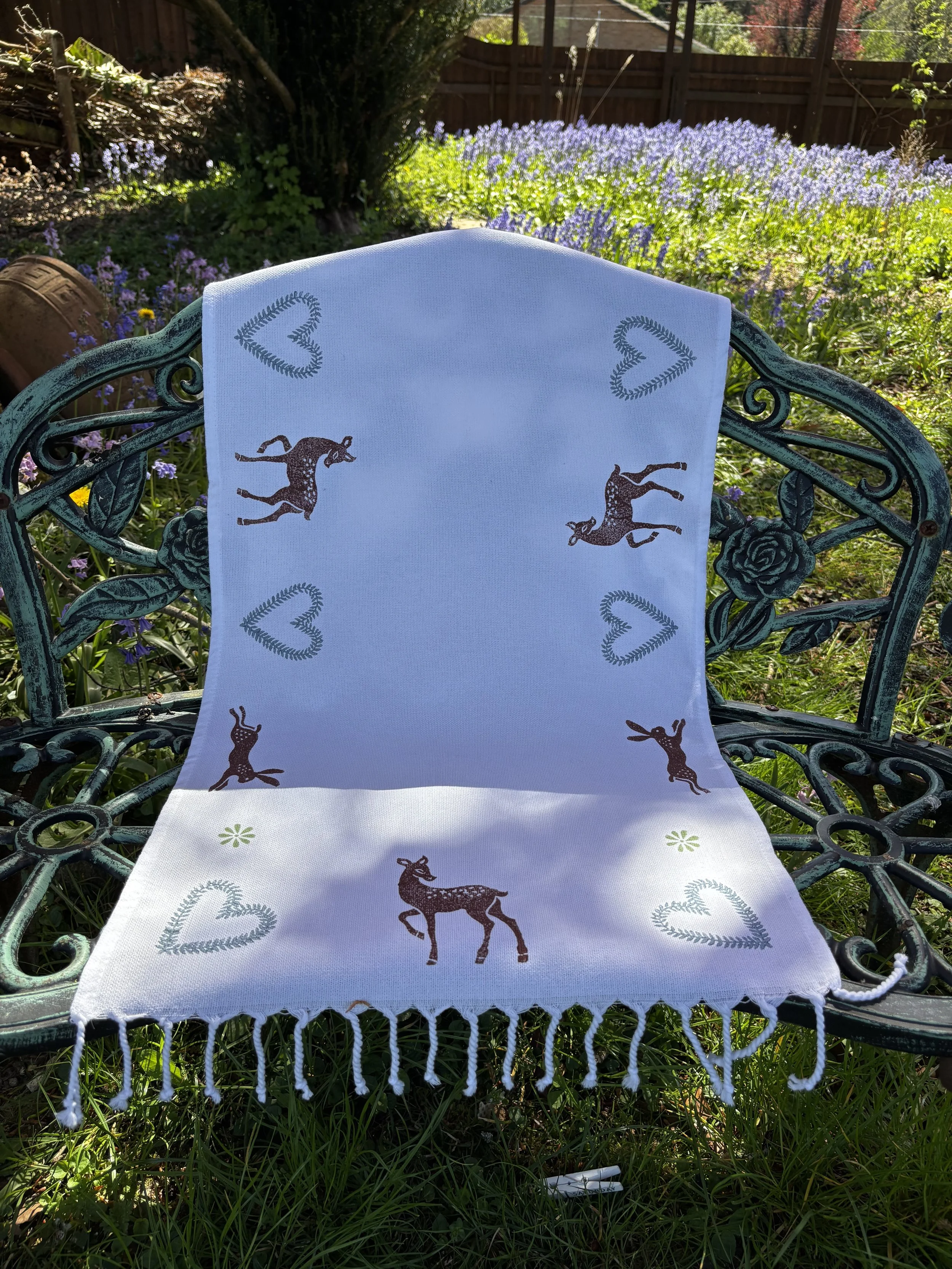 A green metal bench with decorative floral patterns, placed outdoors in a garden. A white table runner with woodblock printed deer and cornflower blue heart patterns drapes over the backrest and seat .