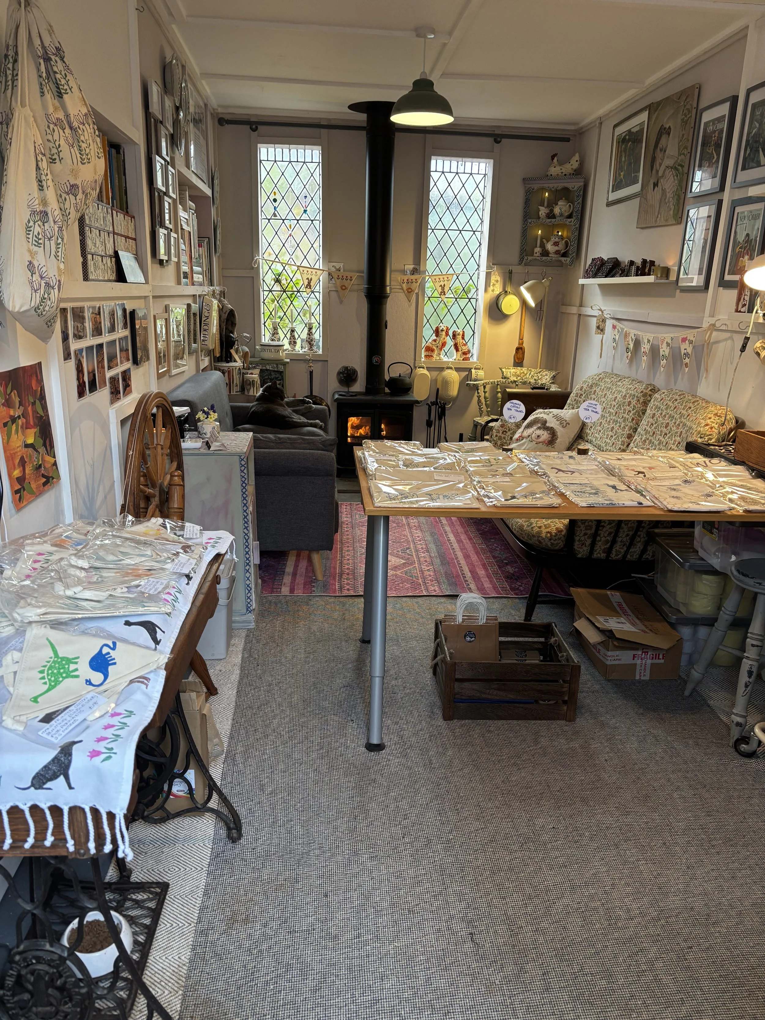 The Hedgepig and Honey studio in winter with thewood stove, a sofa, a coffee table, a bookshelf, and decorated with paintings, photos, and decorative items. There is a cat, Sid Grayson, lying by the wood stove on the sofa