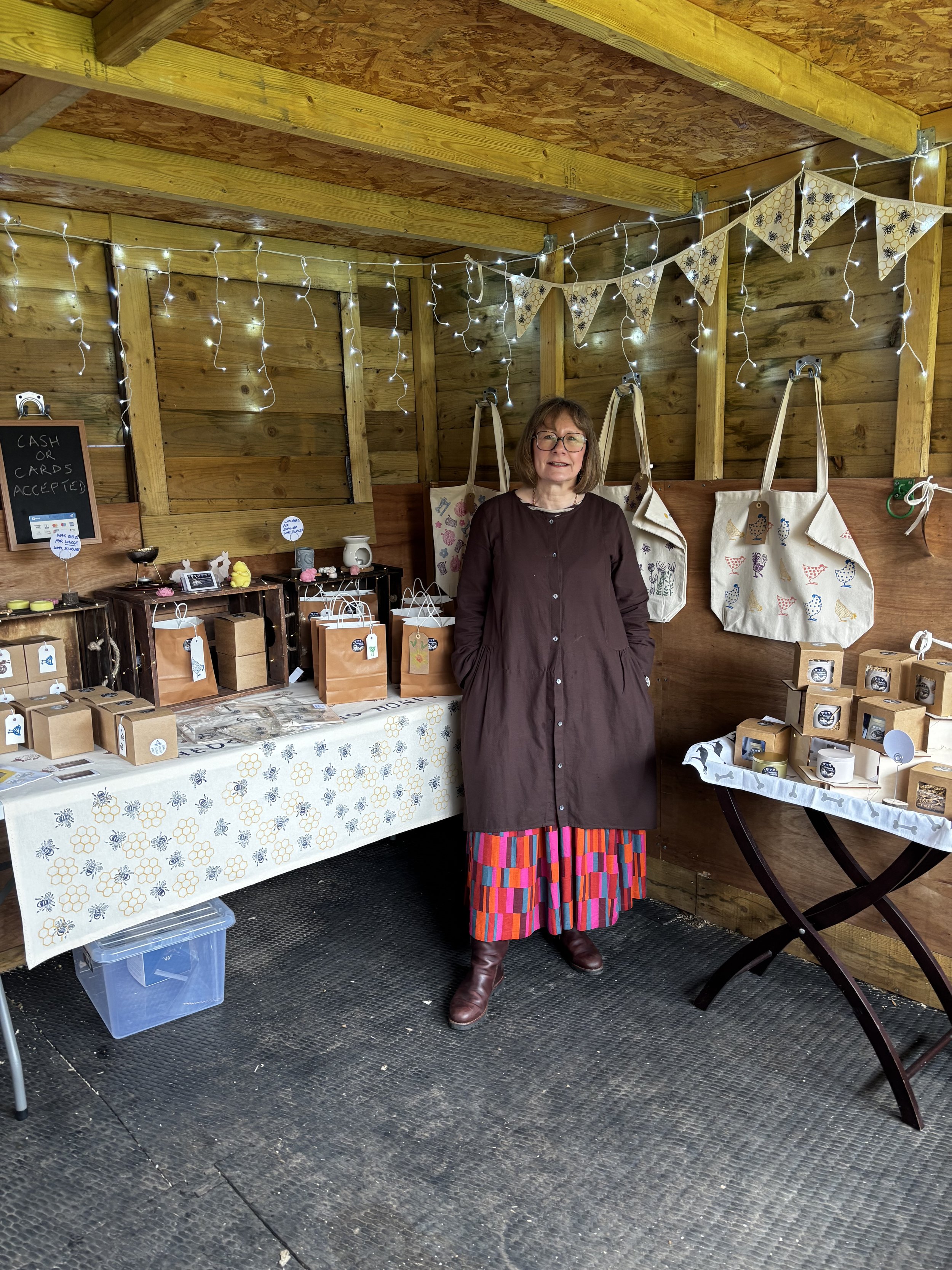 A woman standing behind a booth at a craft fair, with items like bags and candles displayed on tables covered with patterned tablecloths. She is wearing glasses, a long brown coat, a multicolored skirt, and brown boots, inside a wooden structure decorated with string lights and pennant banners.