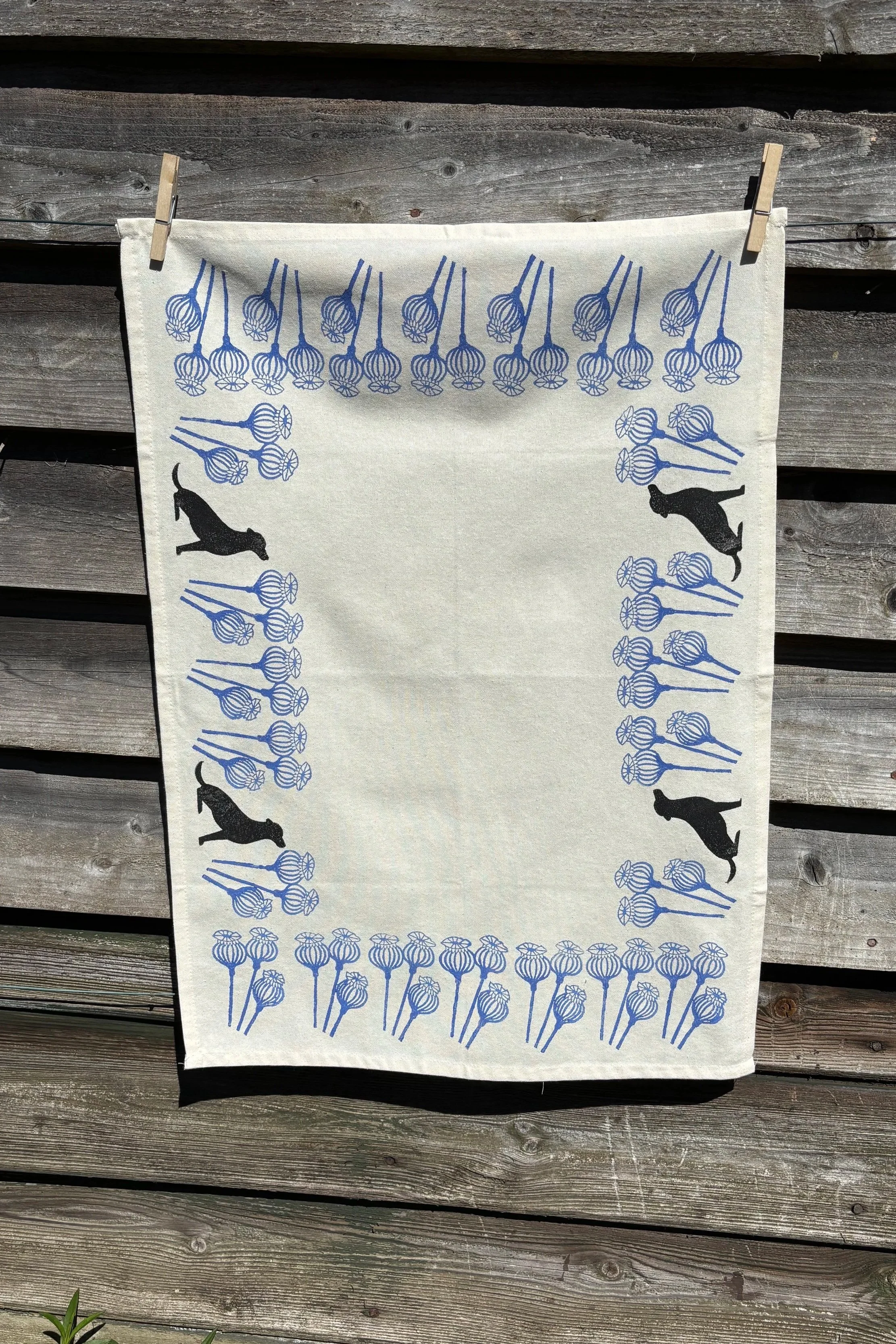 An organic cotton tea towel woodblock printed with cornflower blue flowers  and black Labrador dogs hanging on a country barn.