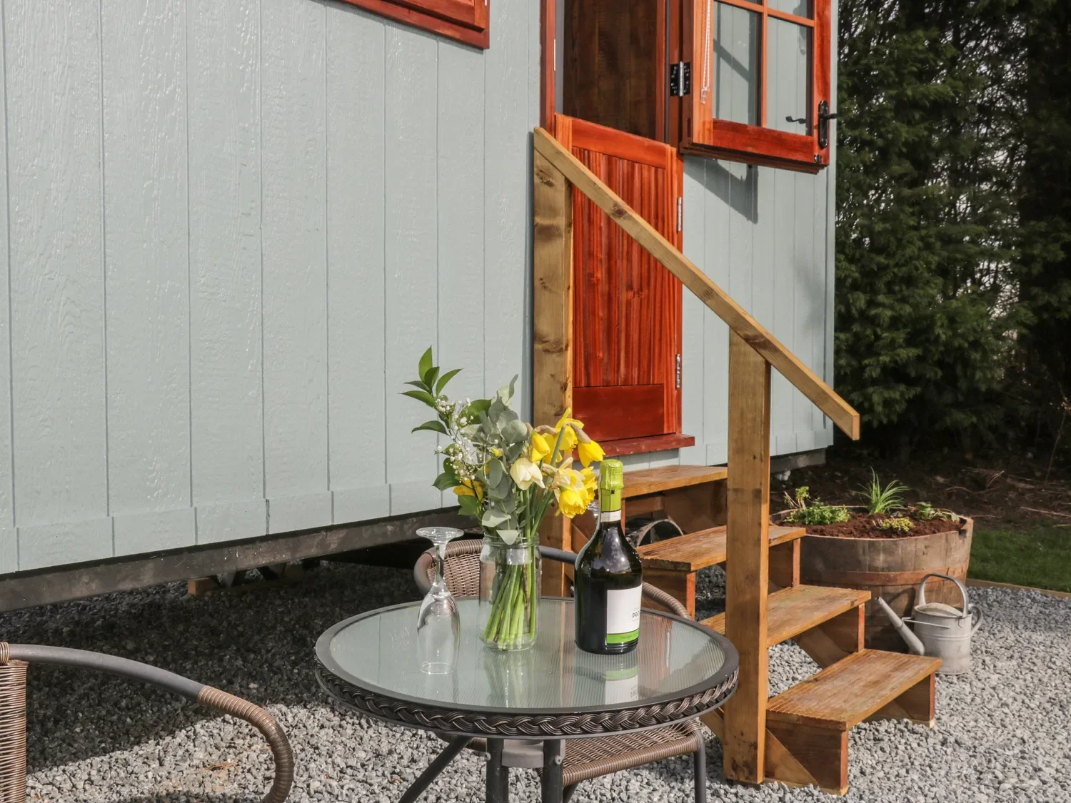 Small outdoor patio with a round glass table holding a vase of flowers and a bottle of wine, adjacent to a tiny staircase leading up to a house with painted wooden siding and open window shutters, surrounded by gravel and plants.