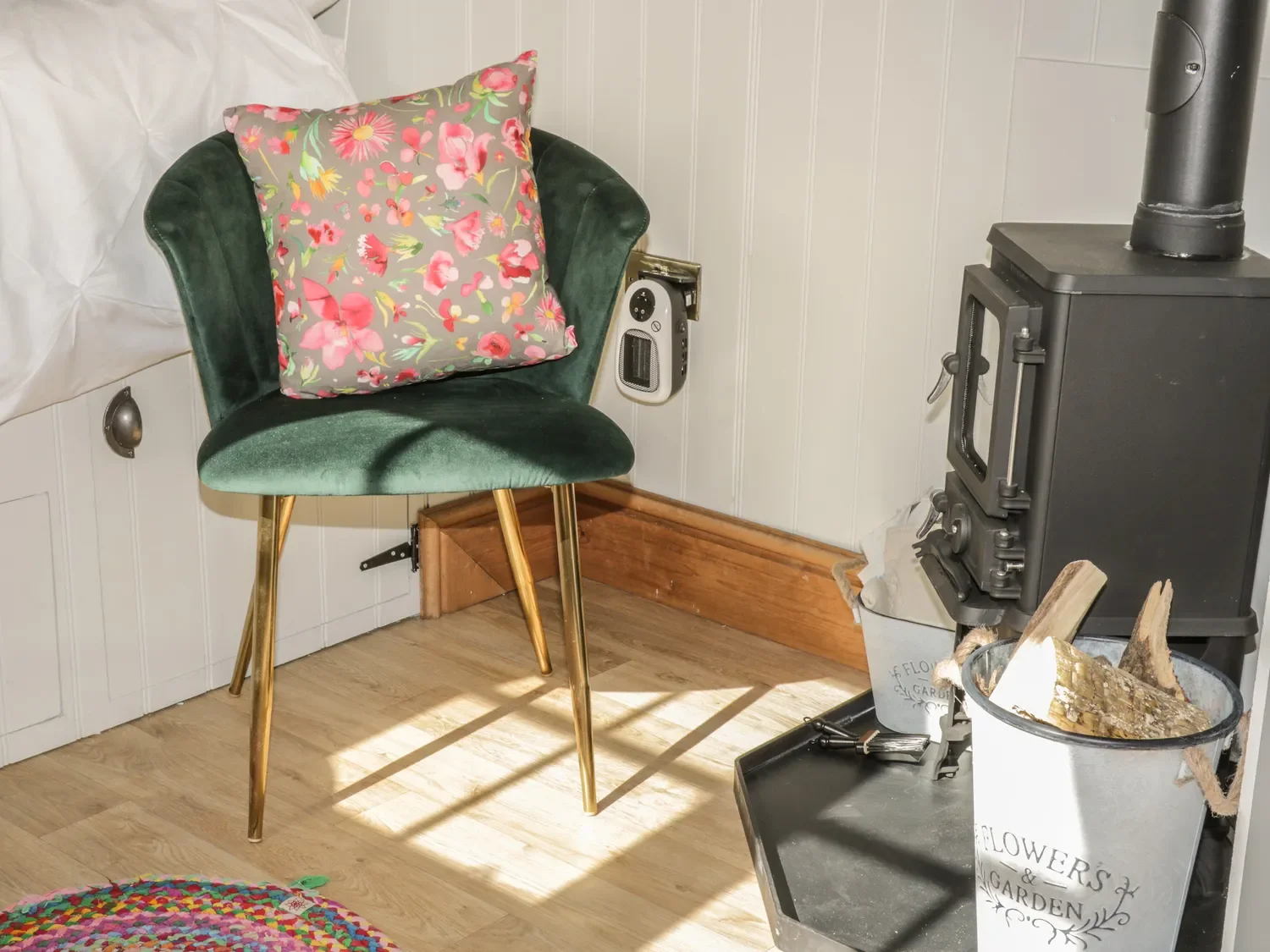 Green velvet chair with gold legs and a floral pillow, next to a white cabinet, a small wall heater, a wood stove with a black pipe, and a bucket of firewood on a black tray.