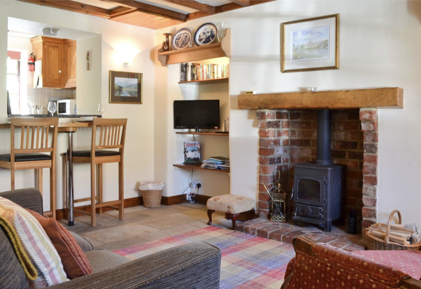 Living room with a wood-burning stove in a brick fireplace, a framed landscape painting on the wall, a bookshelf with books, a small cushioned footstool, a flat-screen TV, and a dining area with high chairs. The room has wooden accents and a colorful
