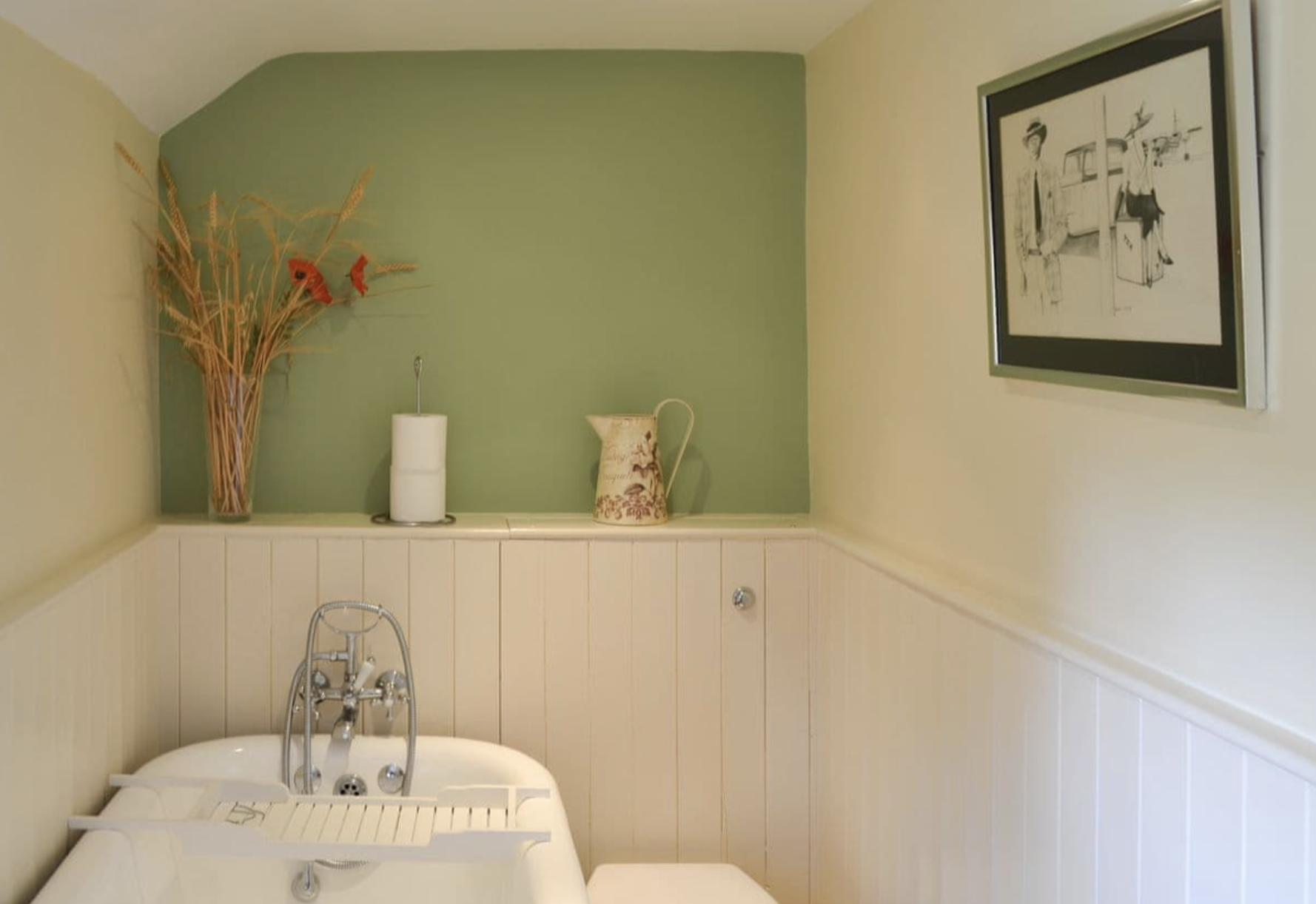 A vintage bathtub with a faucet in a bathroom with beadboard wainscoting and a shelf decorated with dried flowers, a paper towel roll, and a floral pitcher, with framed black and white art on the beige wall.