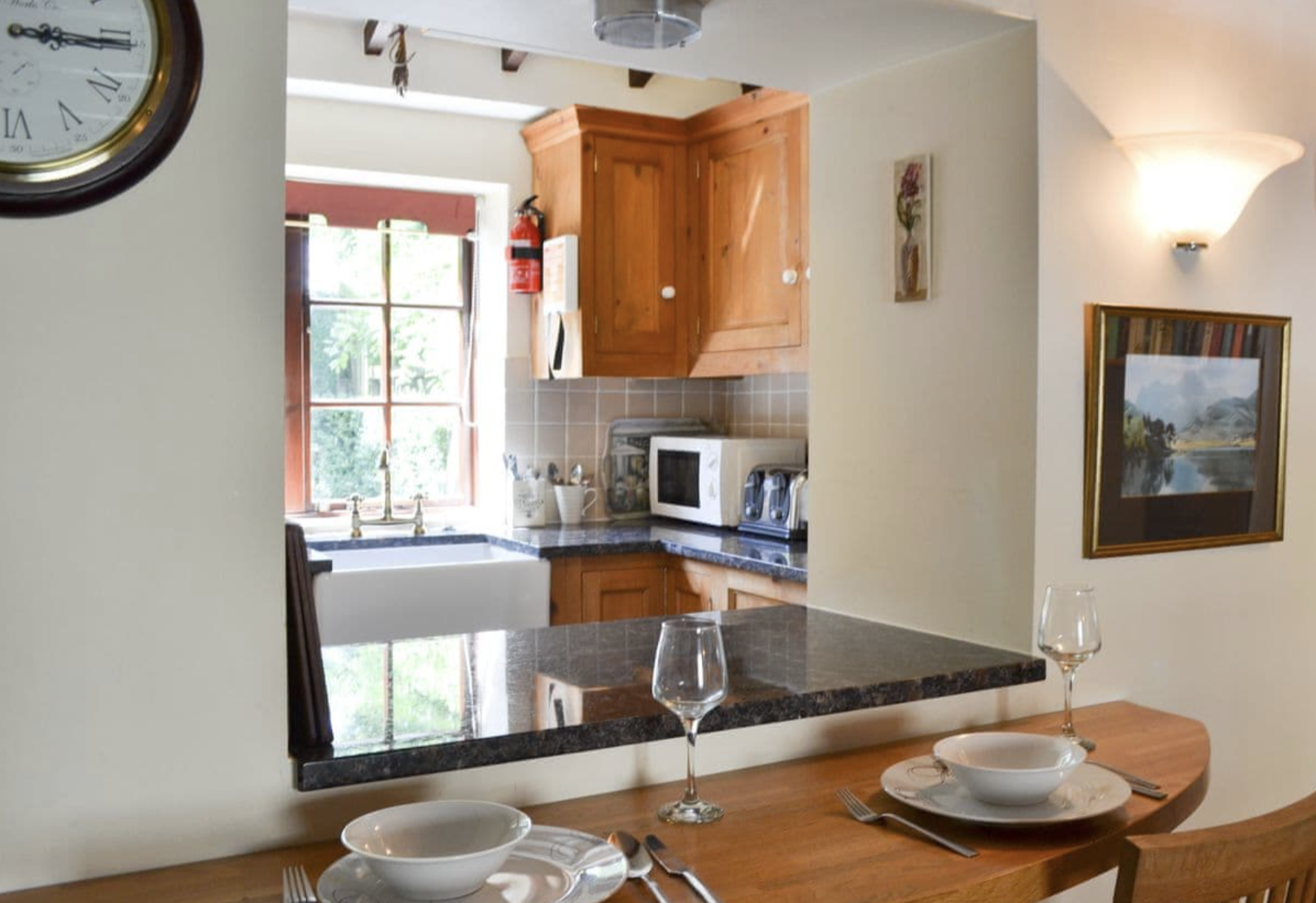 View of a kitchen with wooden cabinets, a window above the sink, a microwave, toaster, and a fire extinguisher on the wall, seen from a dining area with a wooden table set with bowls, glasses, and silverware.