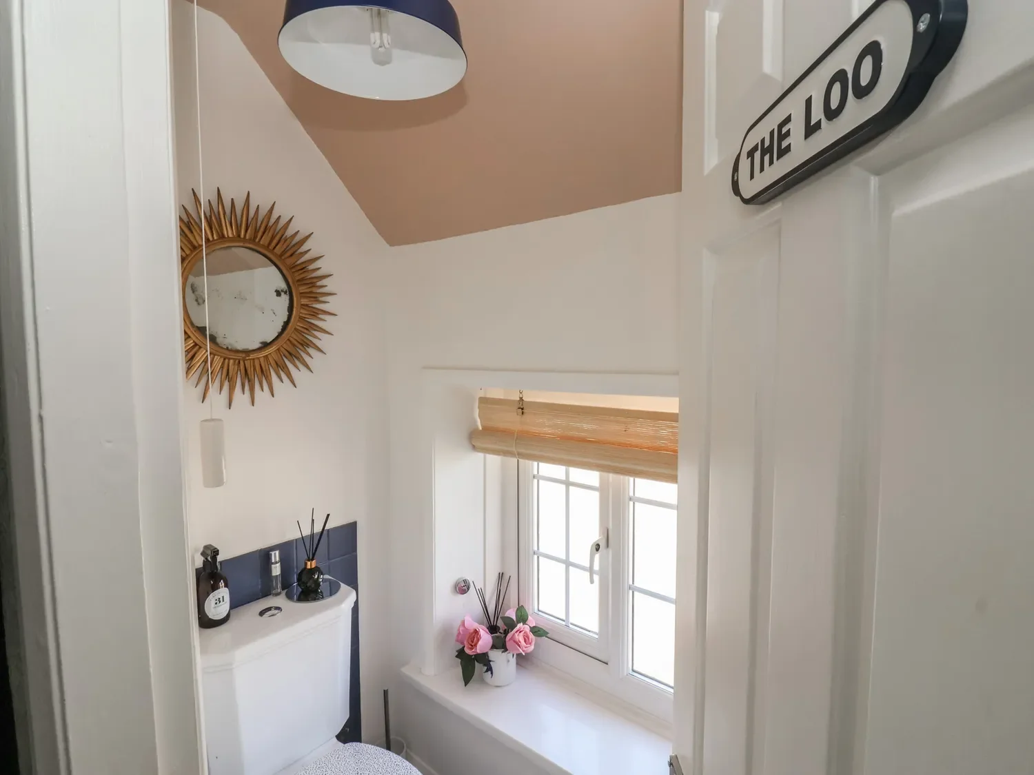 A small bathroom with a white toilet, a window with a beige Roman shade, pink roses in a white vase, a decorative mirror with a sunburst frame, and a wall-mounted sign reading 'The Loo'.