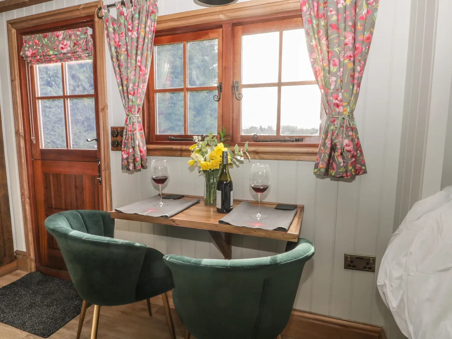 A cozy dining nook with a small wooden table set for two with wine glasses, a wine bottle, and a vase of yellow flowers. Green velvet chairs are on each side, and there are wooden-framed windows with floral curtains and a door with a glass panel, all