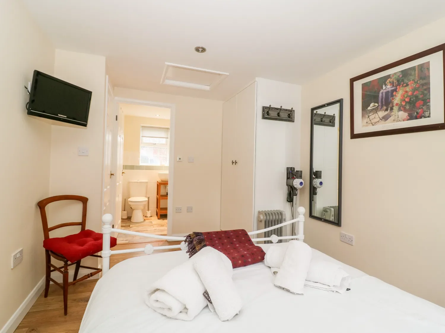 A cozy hotel room with a bed, folded towels, a chair with a red cushion, a small bathroom with a toilet, a wall-mounted TV, a mirror, artwork, and a hairdryer.