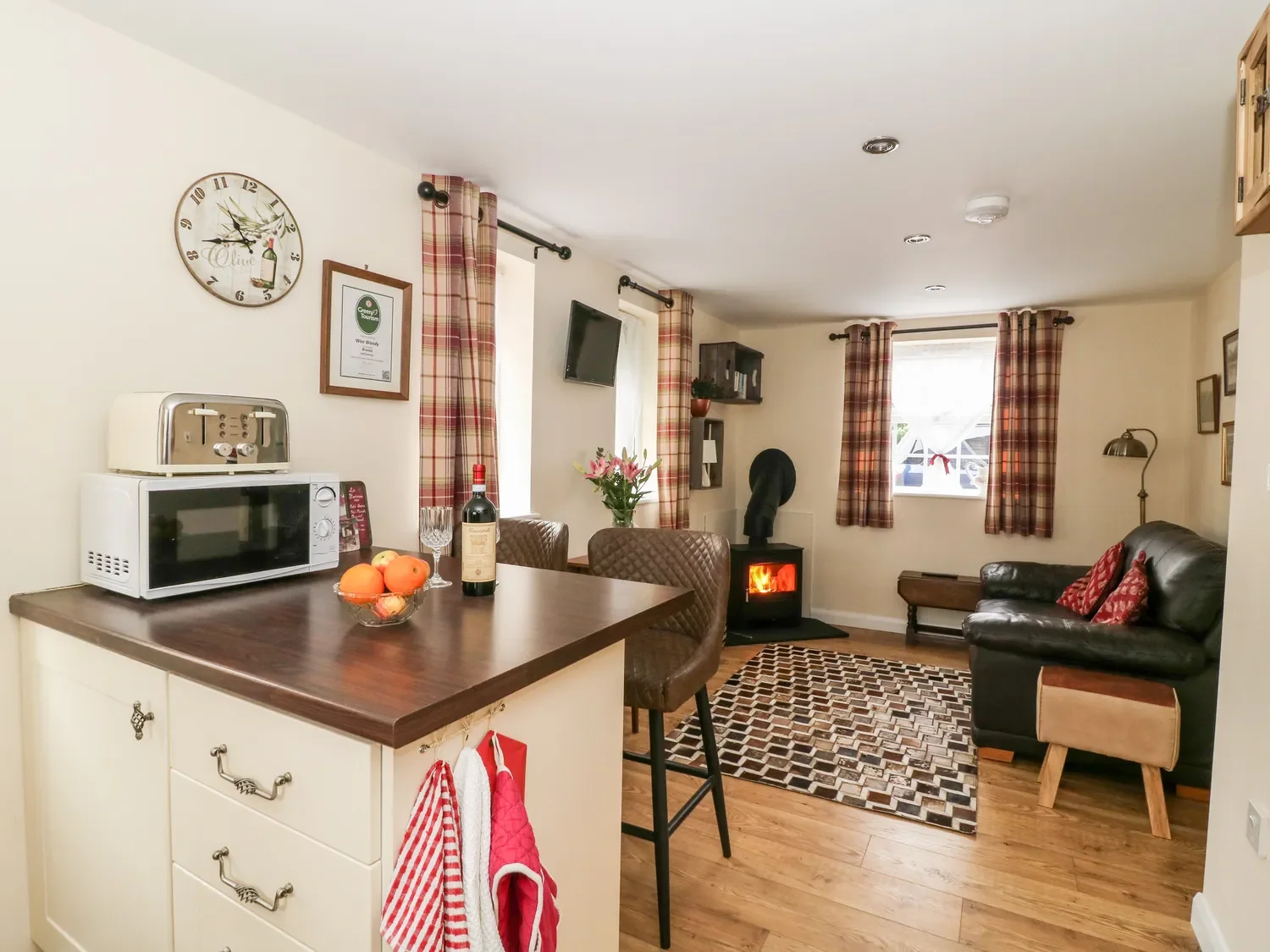 Living room with a window, plaid curtains, a wall clock, wall art, a small TV, a wood stove, a black leather couch, a colorful rug, a dining table with chairs, a bowl of fruit, a bottle of wine, and a microwave on a kitchen counter.