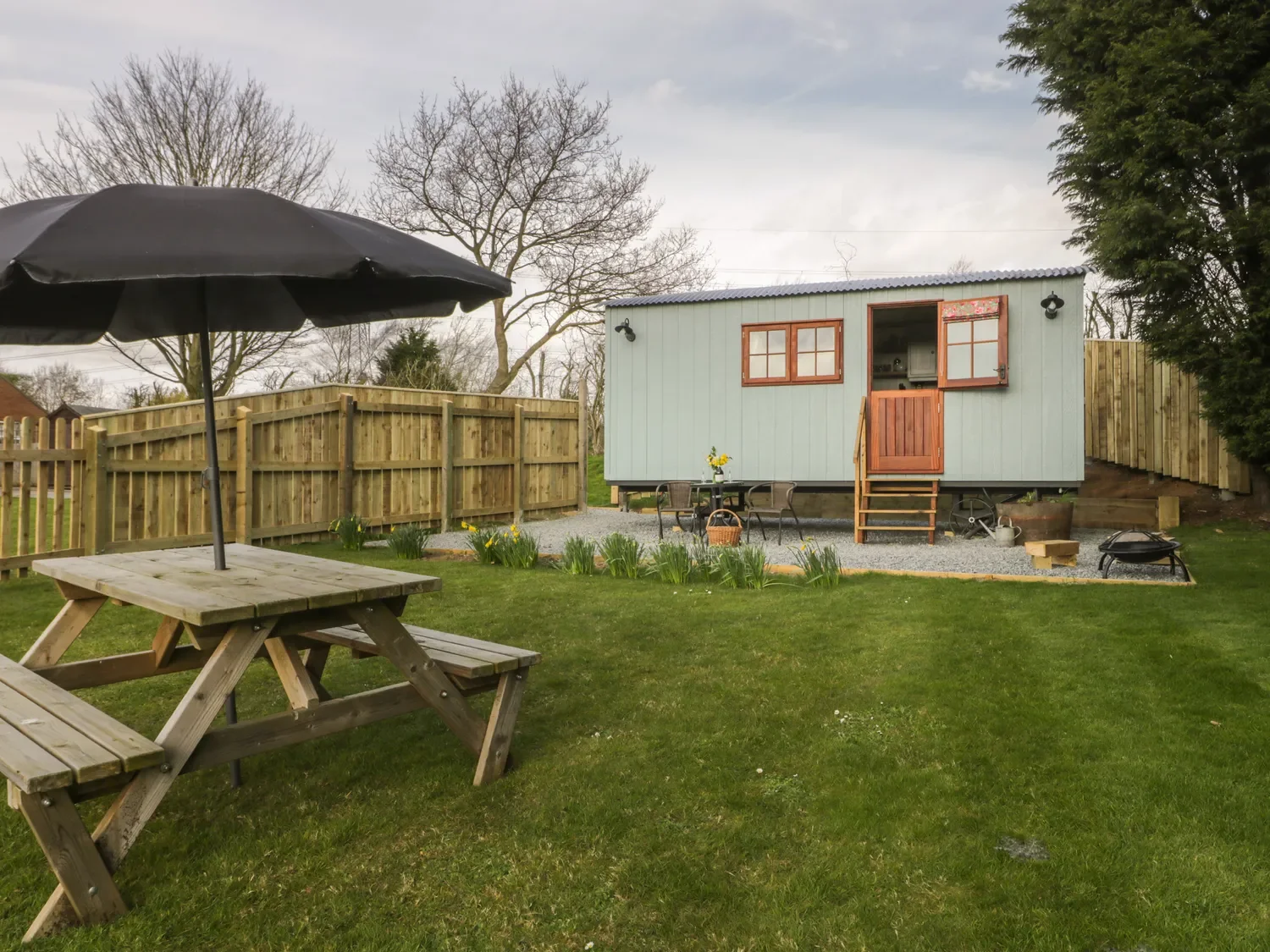 Backyard with a small house or cabin, outdoor dining table with chairs, picnic table with large umbrella, wooden fence, grass lawn, trees, and gardening plants.