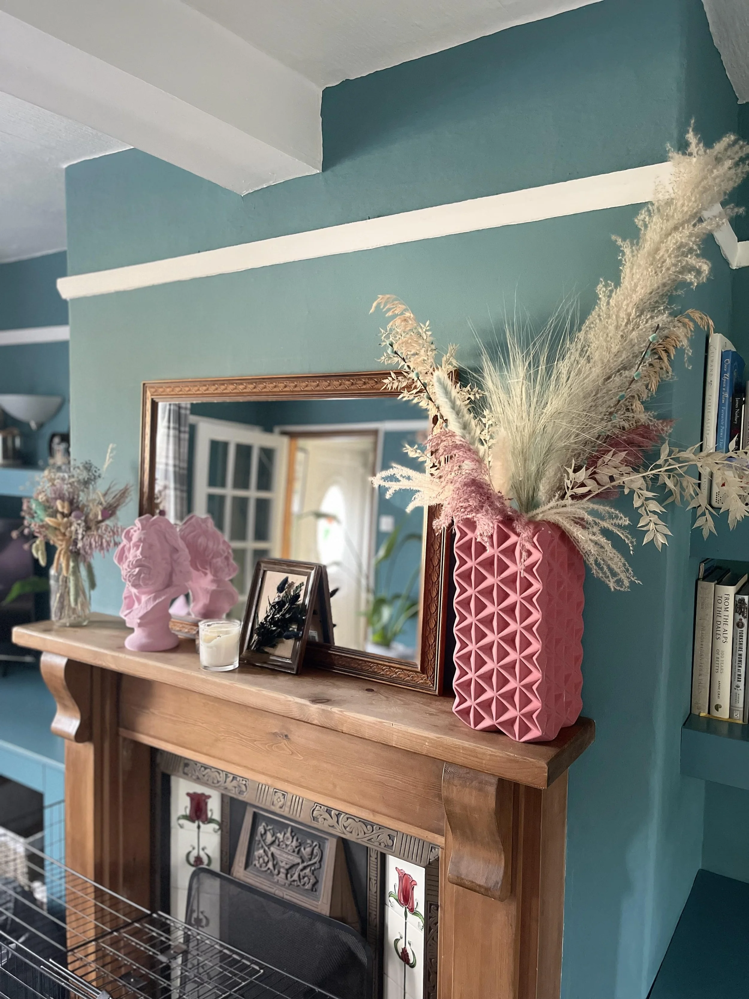 A wooden console table with framed artwork, a pink vase with dried floral arrangements, a pink decorative bust, and a candle. A large rectangular mirror hangs above the table, reflecting a door and a window with curtains. The background wall is painted teal.