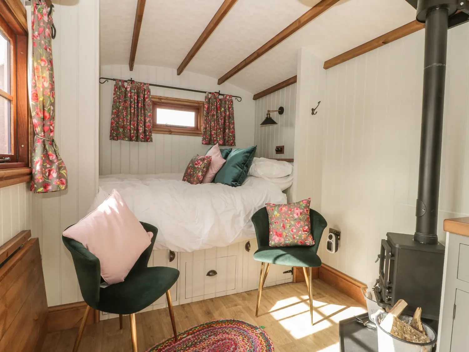 Cozy bedroom with a bed elevated on storage drawers, decorated with floral pillows and curtains, two green chairs with pink pillows, a small window, wood floors, wood beams on the ceiling, and a small stove in the corner.