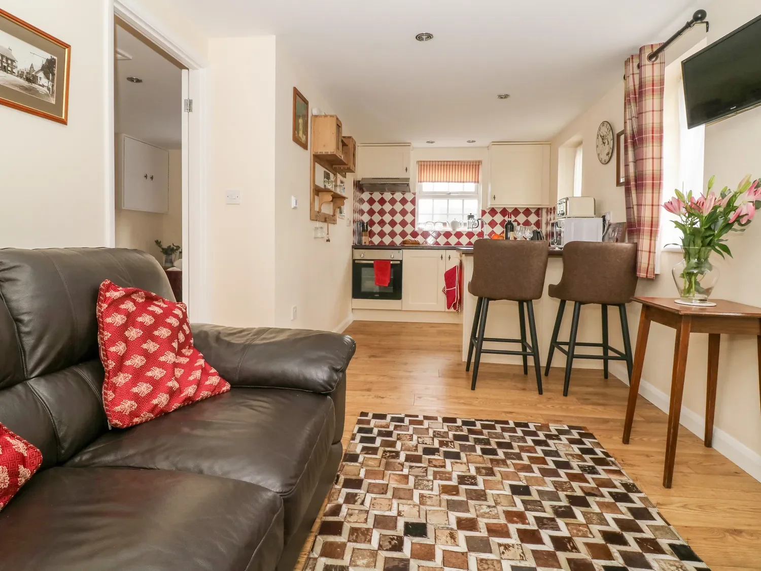 Living room with a brown leather sofa, red patterned pillows, a geometric rug, and an open kitchen with bar stools, checkered backsplash, and window with red plaid curtains.