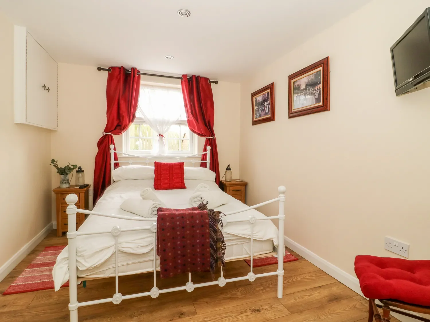 Bedroom with white metal bed frame, red curtains, and wooden nightstands, decorated with red accents and framed pictures on the wall.