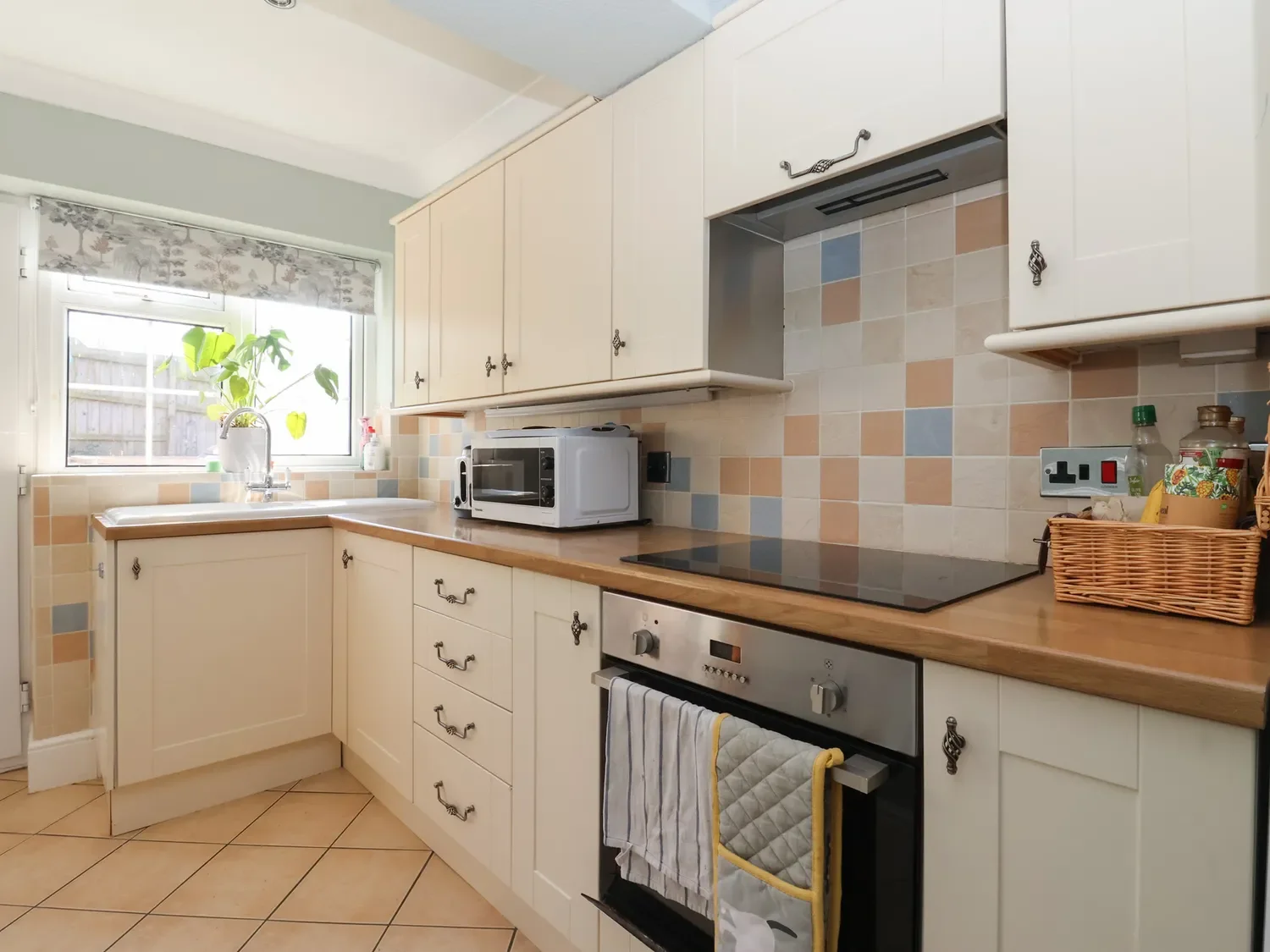 Kitchen with cream cabinets, beige tiled backsplash, black electric stove, microwave, and a window with a plant on the windowsill