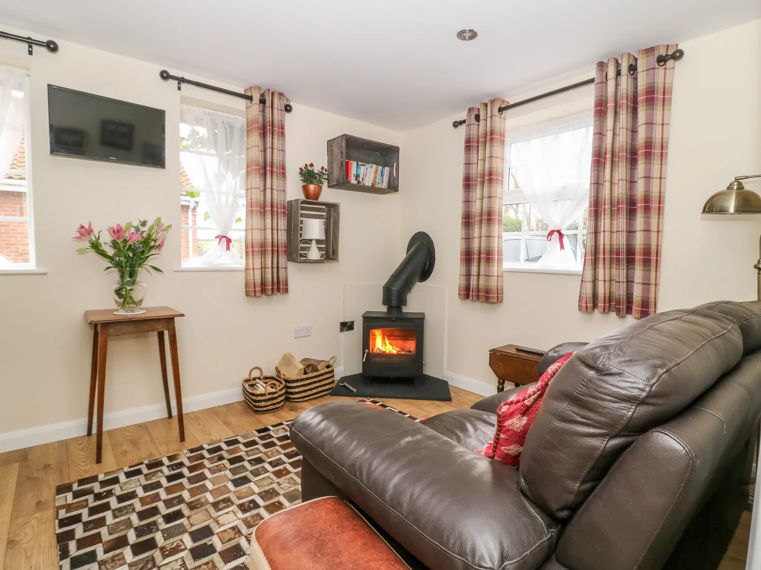 Cozy living room with a leather sofa, wood floors, a small wood stove with visible fire, framed windows with plaid curtains, a hanging TV, a small side table with flowers, and decorative baskets.