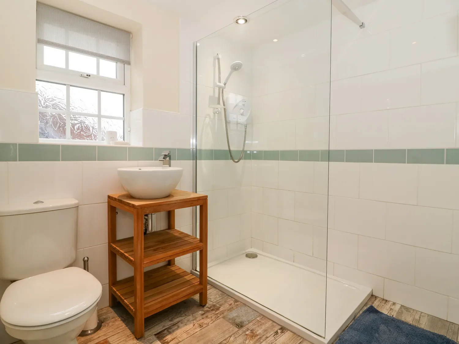 Bathroom with a shower area enclosed by glass, a toilet, a wooden stand with a vessel sink, a window with blinds, wooden flooring, and a blue rug.