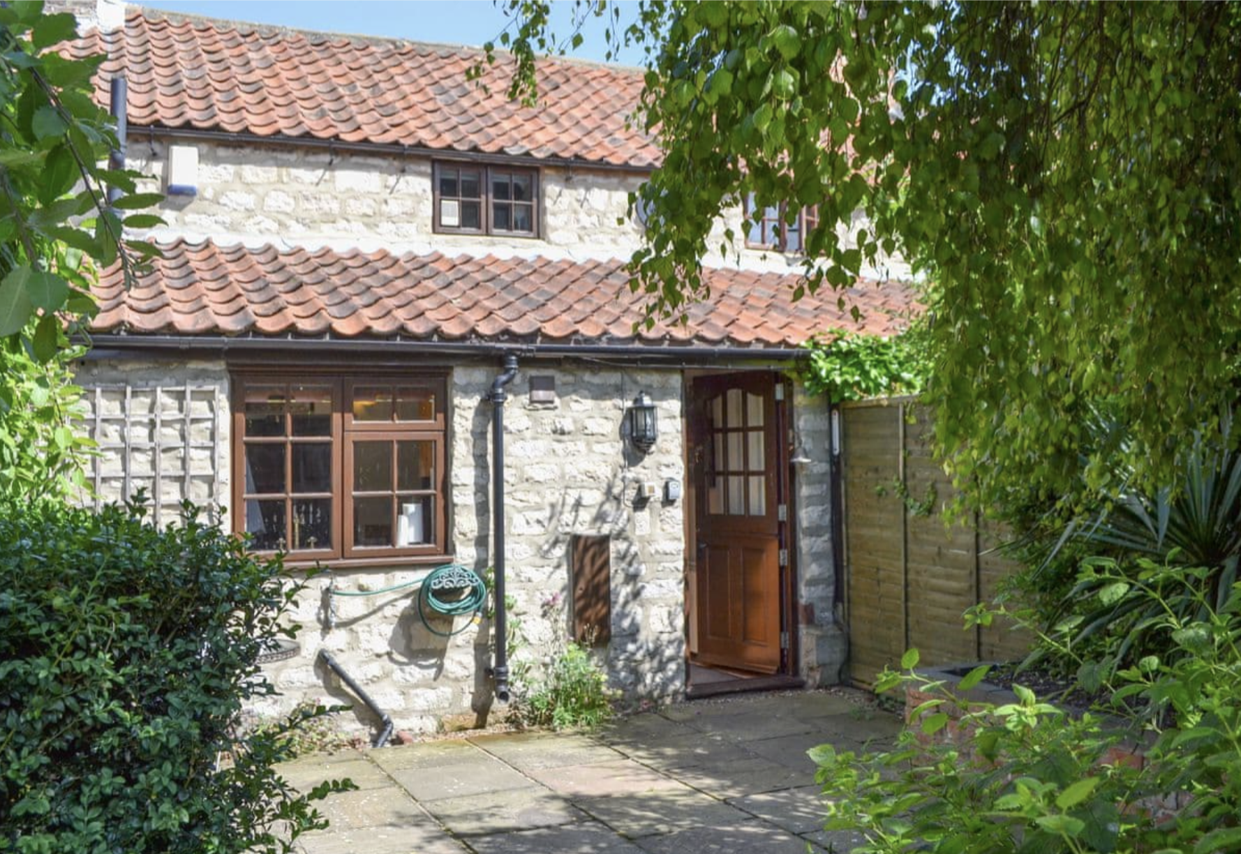 A charming stone cottage with a red-tiled roof, surrounded by lush green plants and trees, featuring a wooden door and window with a grid pattern.