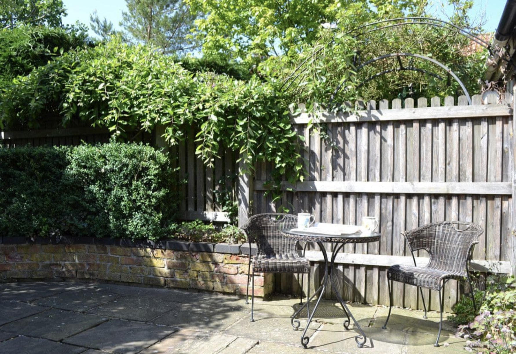 A cozy backyard patio with a round metal table and three matching chairs, surrounded by green bushes, potted plants, and a wooden fence, under a clear blue sky.