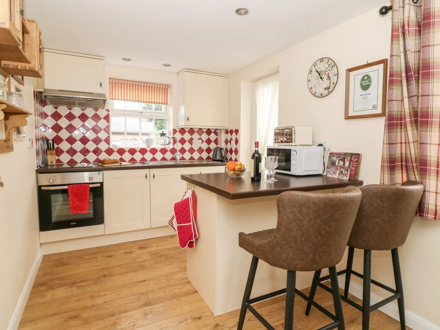 Cozy kitchen with red and white checkered backsplash, white cabinets, wooden countertops, and wooden flooring. Includes small appliances and table with two chairs.