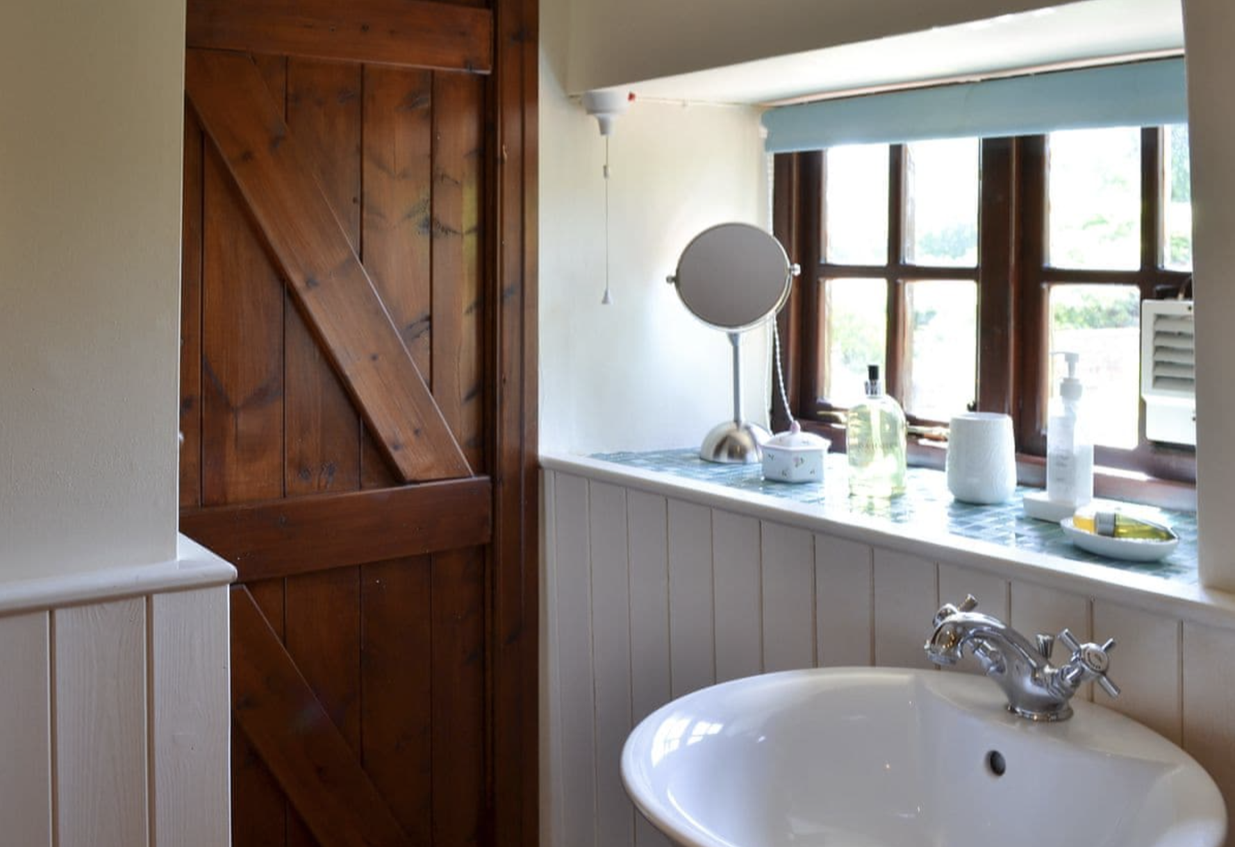 A bathroom windowsill with a small mirror, glass bottles, a soap dispenser, and other toiletries. A white sink with a chrome faucet is in the foreground, and a closed wooden door is on the left side of the image.
