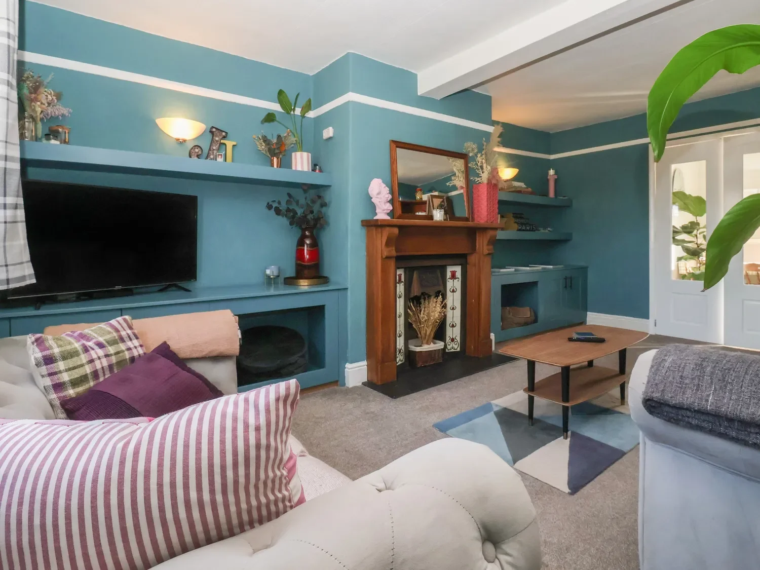 Living room with blue walls, a white and pink striped pillow on a beige sofa, a small wooden coffee table, a TV on a blue built-in shelf, a fireplace with a wooden mantel, and various decorative items including vases, plants, and framed pictures.