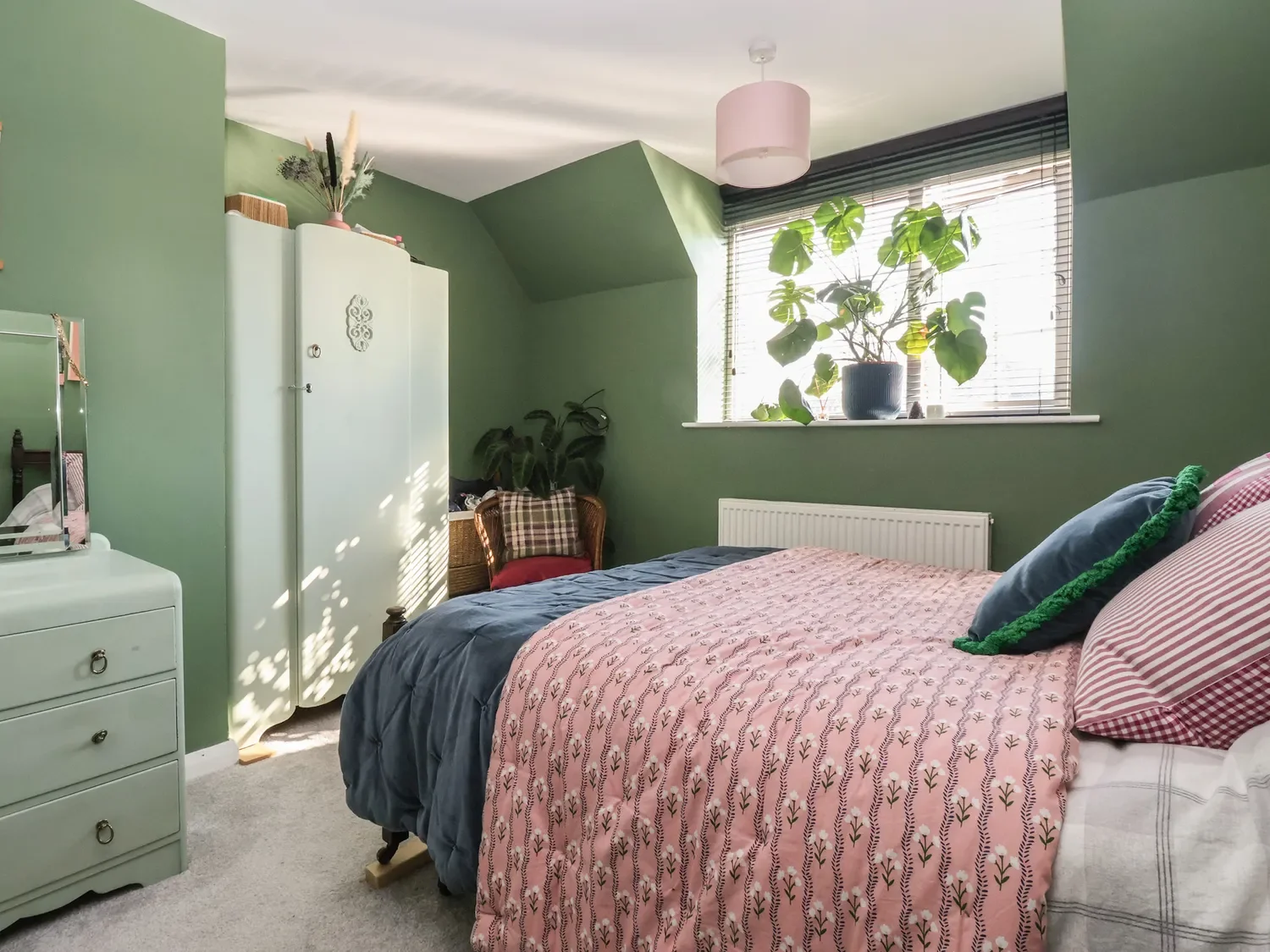 A bedroom with green walls, a bed with pink and navy bedding, a window with blinds, and various plants on the windowsill.