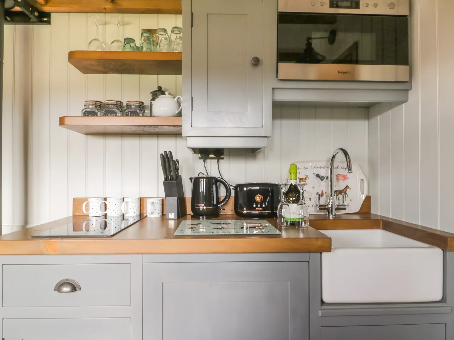 A compact kitchen with a wooden countertop, open shelves with glasses, jars labeled for coffee, tea, and sugar, a white teapot, a black electric kettle, a toaster, and a bottle of wine. There are four coffee mugs with polka dots, a knife block with k