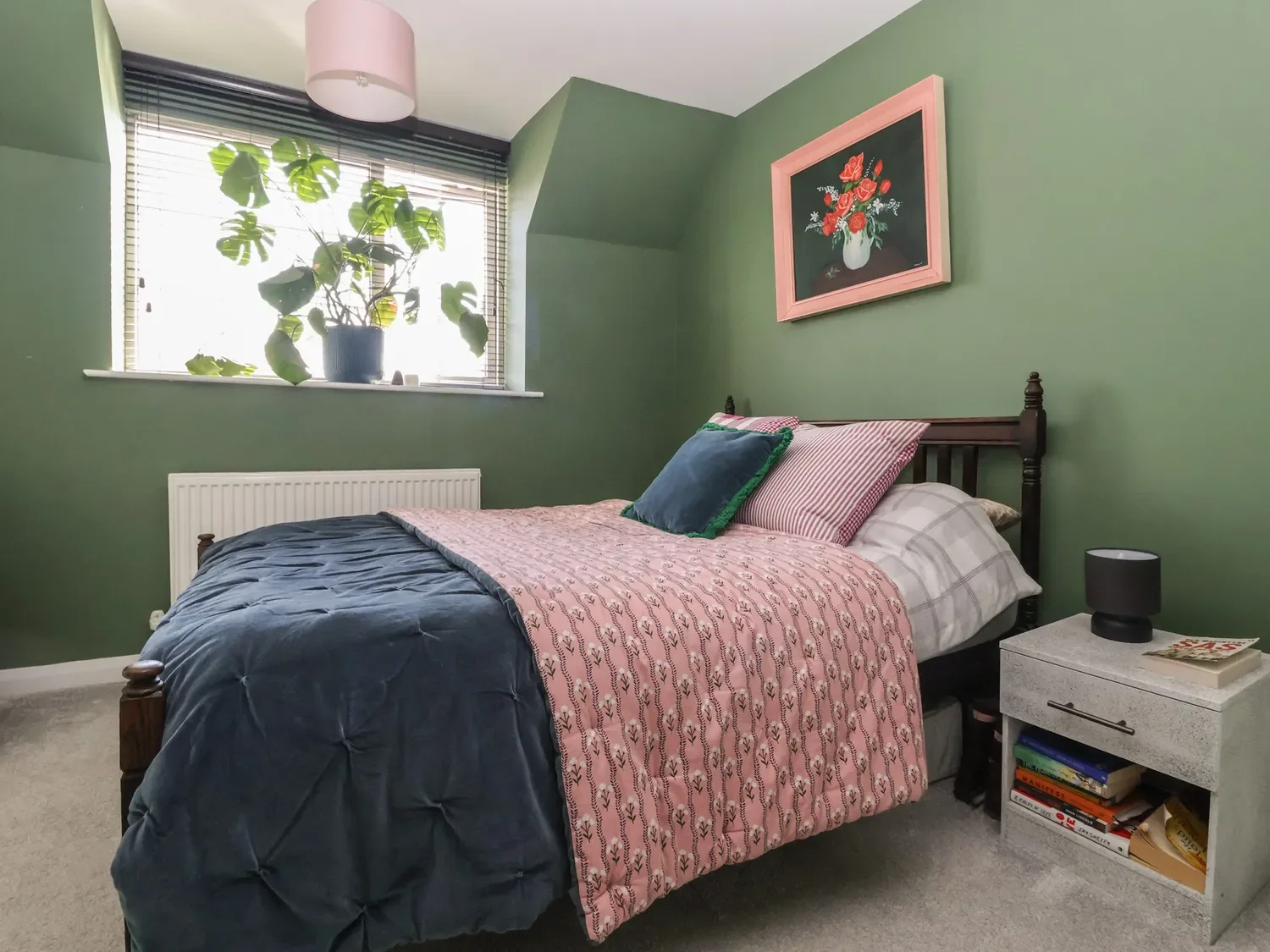 A cozy bedroom with green walls, a window with blinds, and a potted plant on the windowsill. The bed has a dark blue comforter, pink patterned blanket, and multiple pillows. There is a nightstand with a black lamp, books, and a magazine. A framed flo