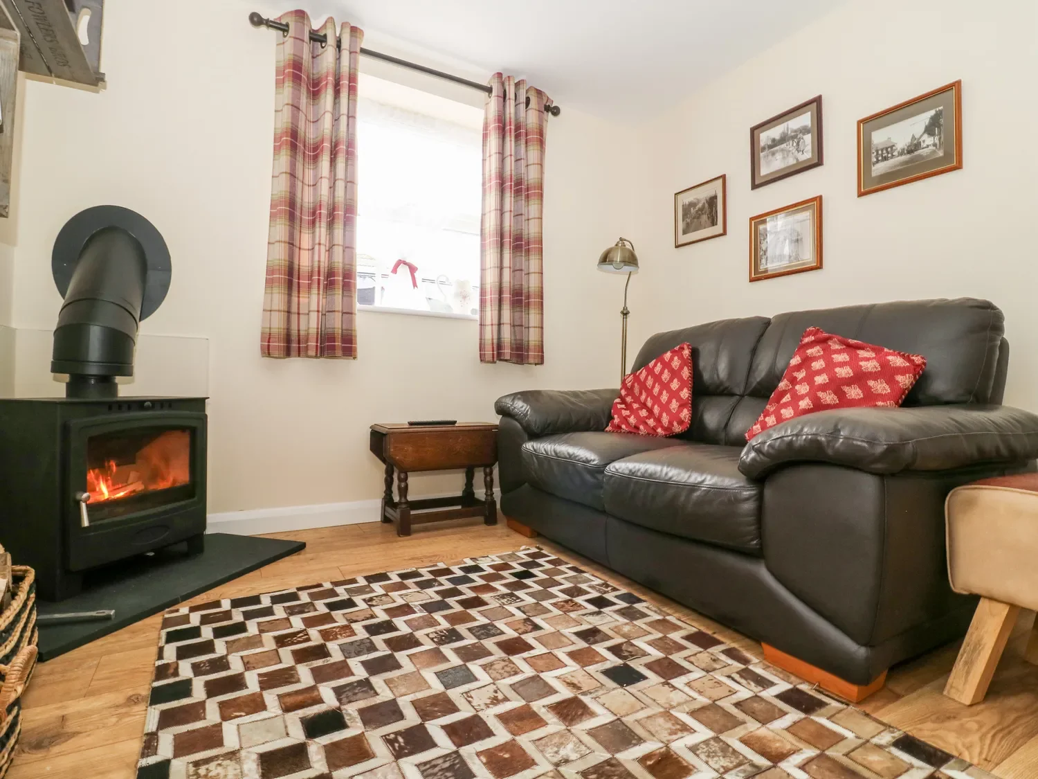 Cozy living room with a black leather sofa, red and white patterned pillows, a wood stove with a fire burning, a small wooden side table, framed black and white photographs on the wall, a window with plaid curtains, a floor lamp, and a patterned area