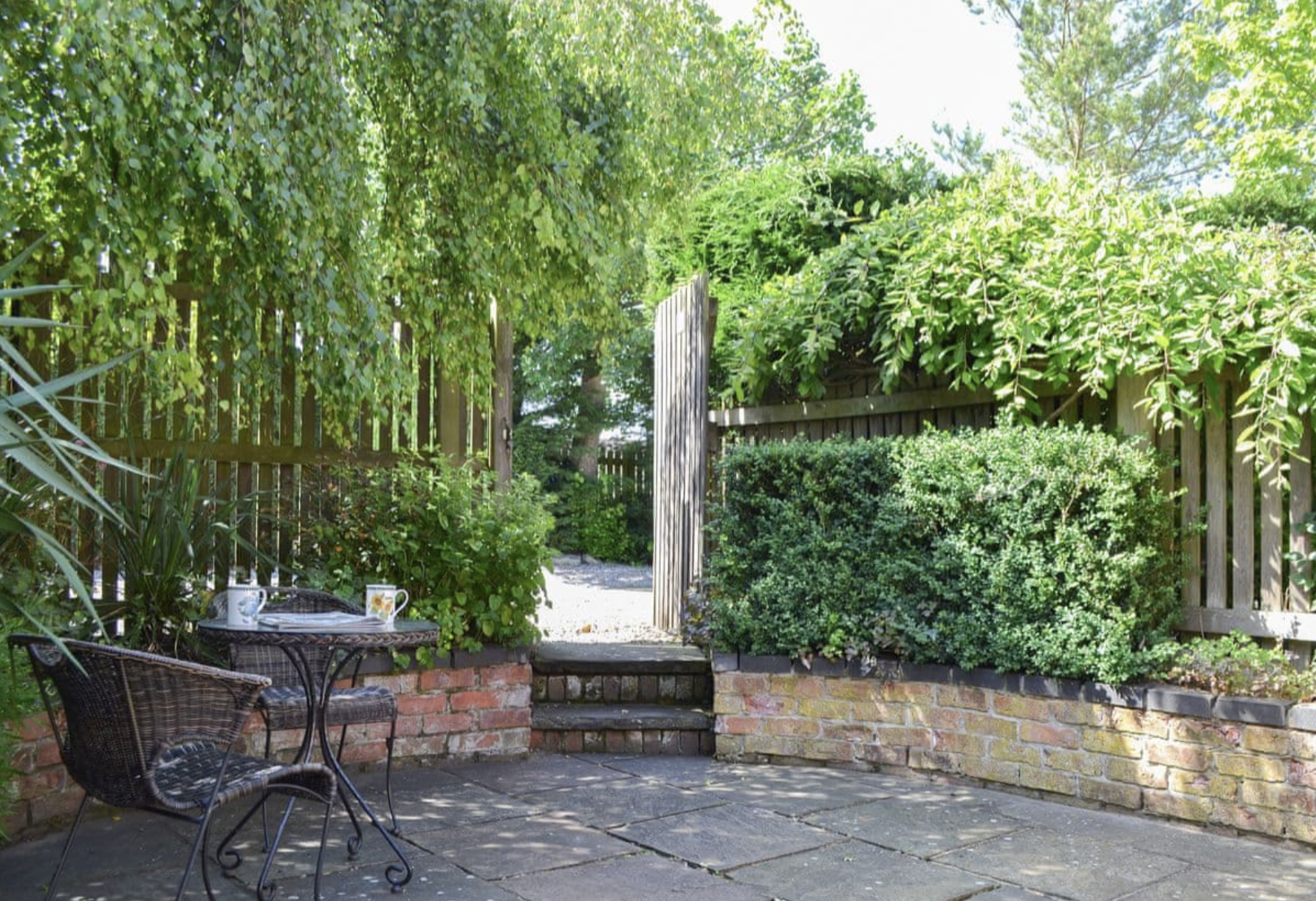 A lush backyard garden featuring a patio with wicker chairs and a small wrought iron table with cups and a book, surrounded by dense greenery, a wooden fence, and a gate leading to a sunny outdoor area.