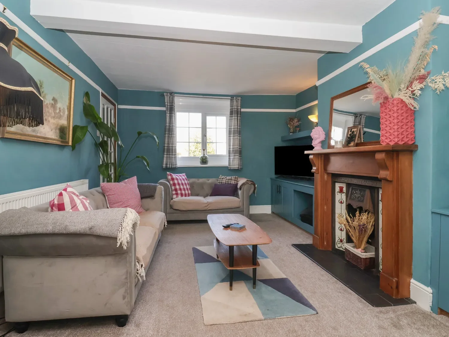 Living room with blue walls, beige sofas with red and white cushions, wooden coffee table, fireplace with a wooden mantel, television, window with plaid curtains, and decorative plants.