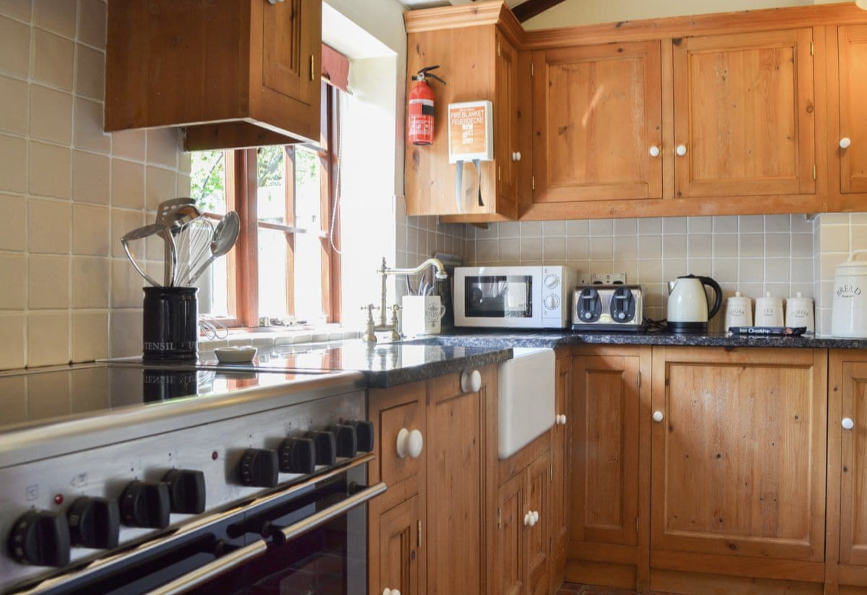 A kitchen with wooden cabinets and a black countertop. There is a window with sunlight, a microwave, a toaster, an electric kettle, and jars with labels on the counter. A fire extinguisher and an emergency fire blanket are mounted on the wall.