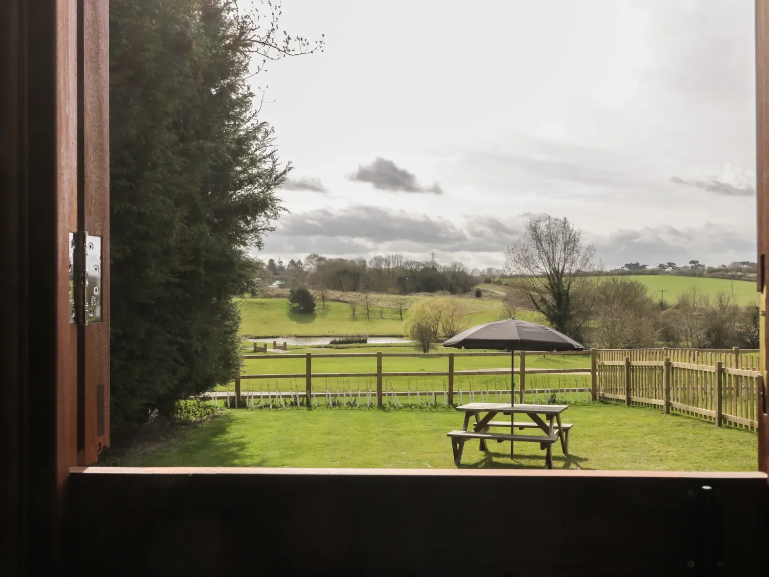 View of a grassy backyard with a picnic table and umbrella, overlooking a lake and rolling green hills with trees under cloudy sky, seen from an open window.
