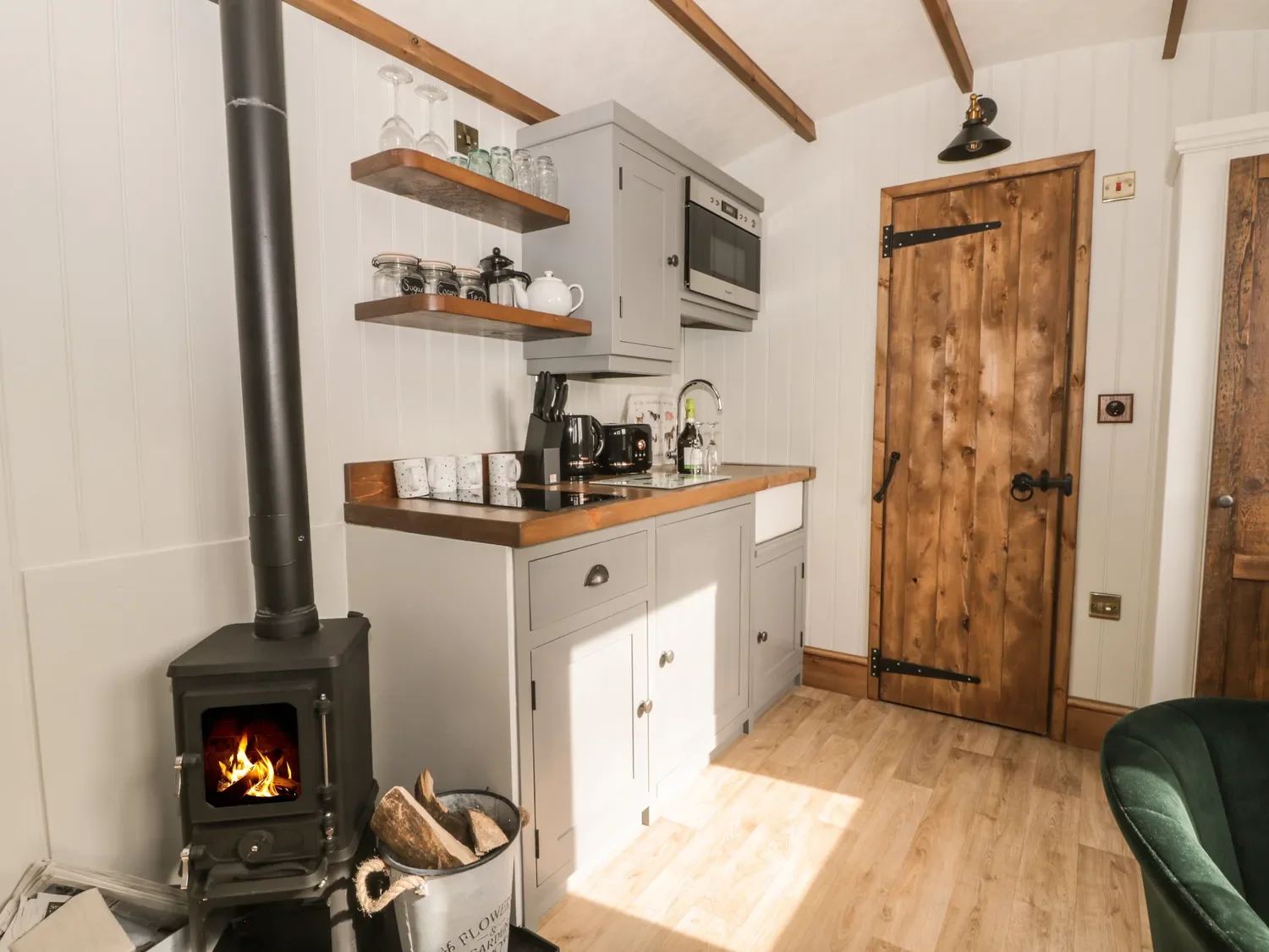 A cozy kitchen with a wood stove emitting a small fire, gray cabinets with wooden countertops, open shelves with glasses and jars, a small sink, coffee machine, and cups, a rustic wooden door, and a green armchair in the corner.