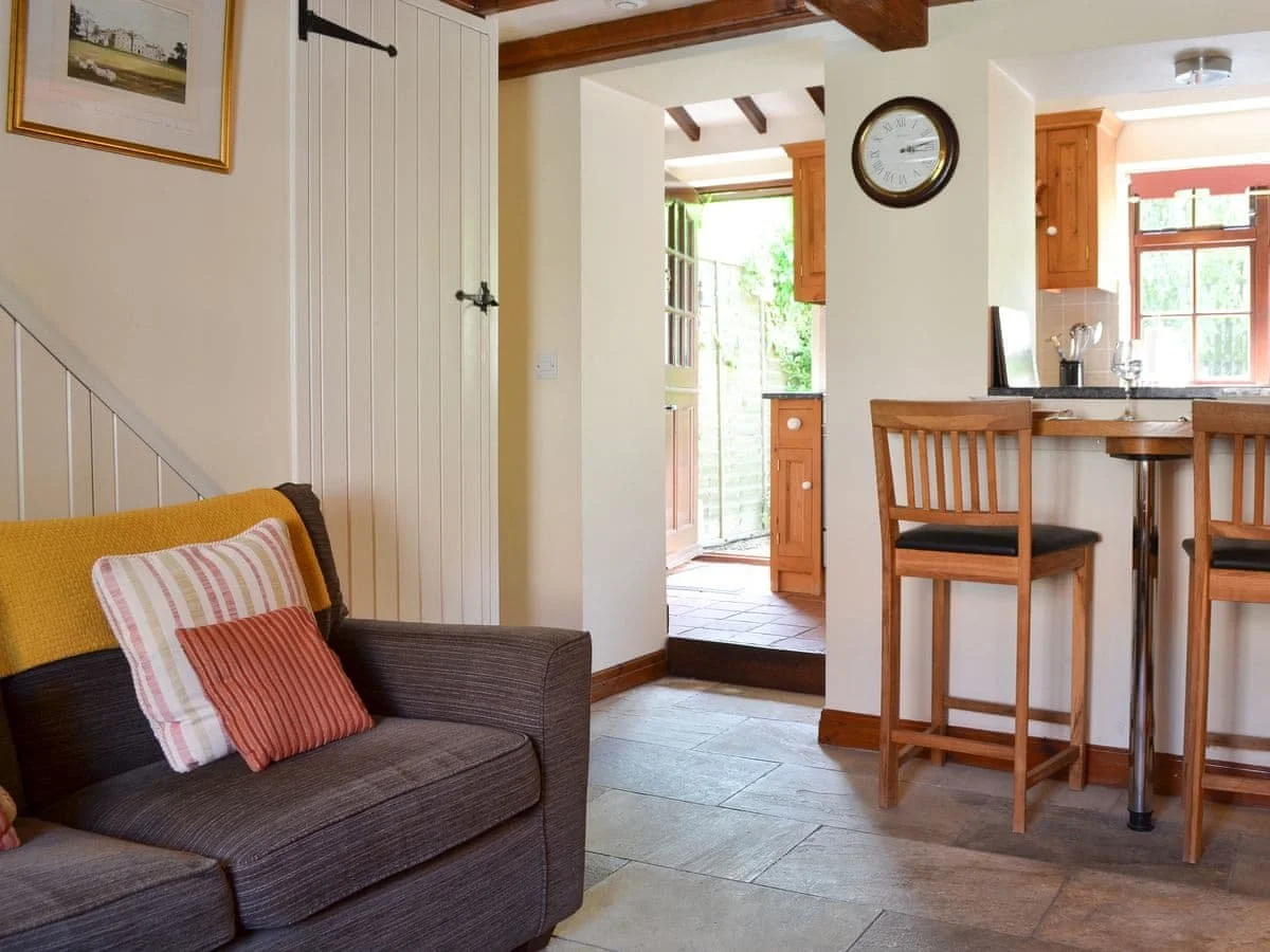 Living room with a brown sofa, colorful cushions, a wall clock, and view into the kitchen with wooden cabinets and bar stools near the counter.