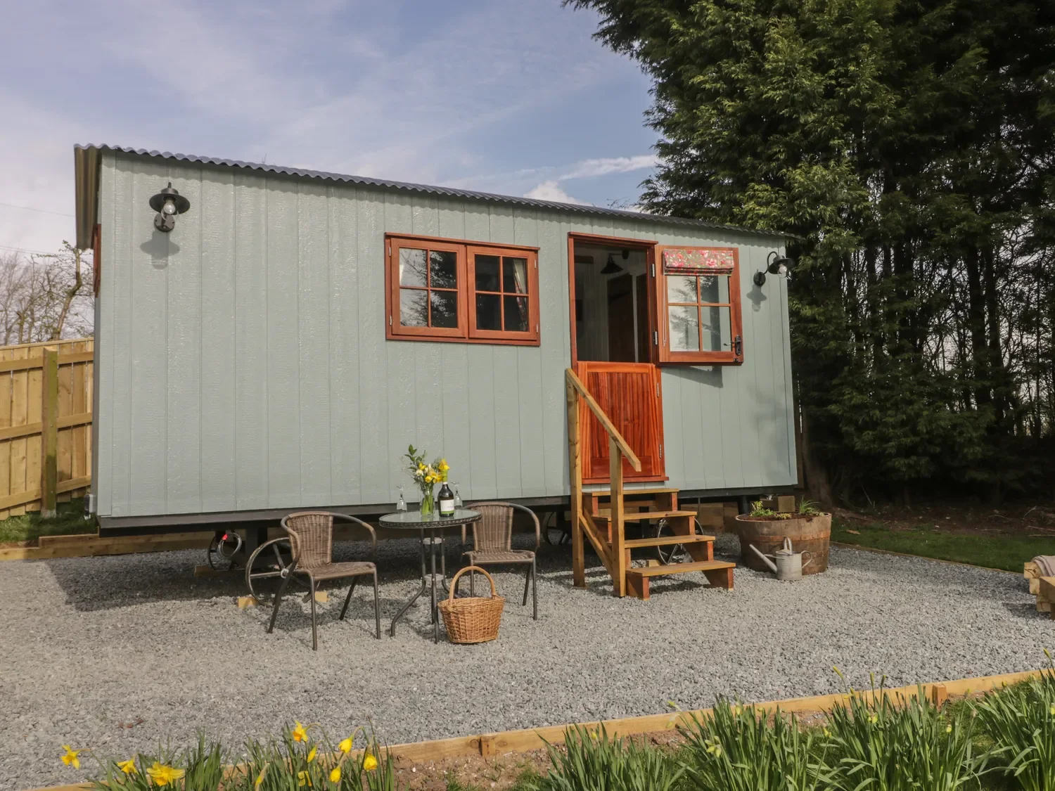 A tiny house with light green exterior walls, wooden window frames, and a small wooden staircase leading to an open door. There is a gravel yard with a small table, two chairs, and a flower arrangement. A wooden fence and trees are in the background.