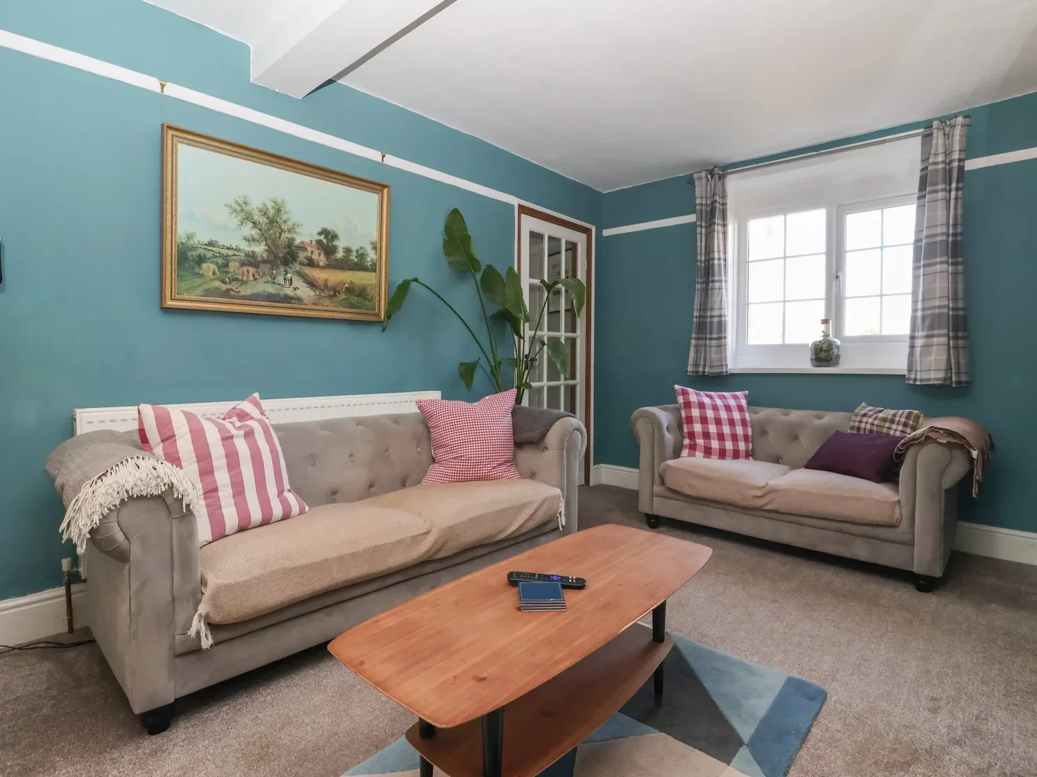 A living room with two beige tufted sofas, pink gingham pillows, a wooden coffee table, a window with plaid curtains, a houseplant, and a landscape painting on teal walls.
