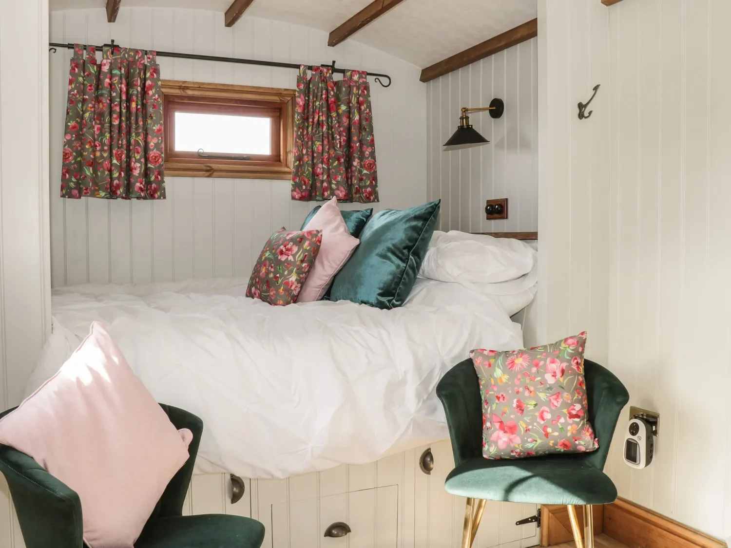 Cozy bedroom with a bed covered in white bedding, topped with decorative pillows, two green velvet chairs with pink pillows, a small window with floral curtains, wood beam ceiling, and a wall-mounted black reading lamp.