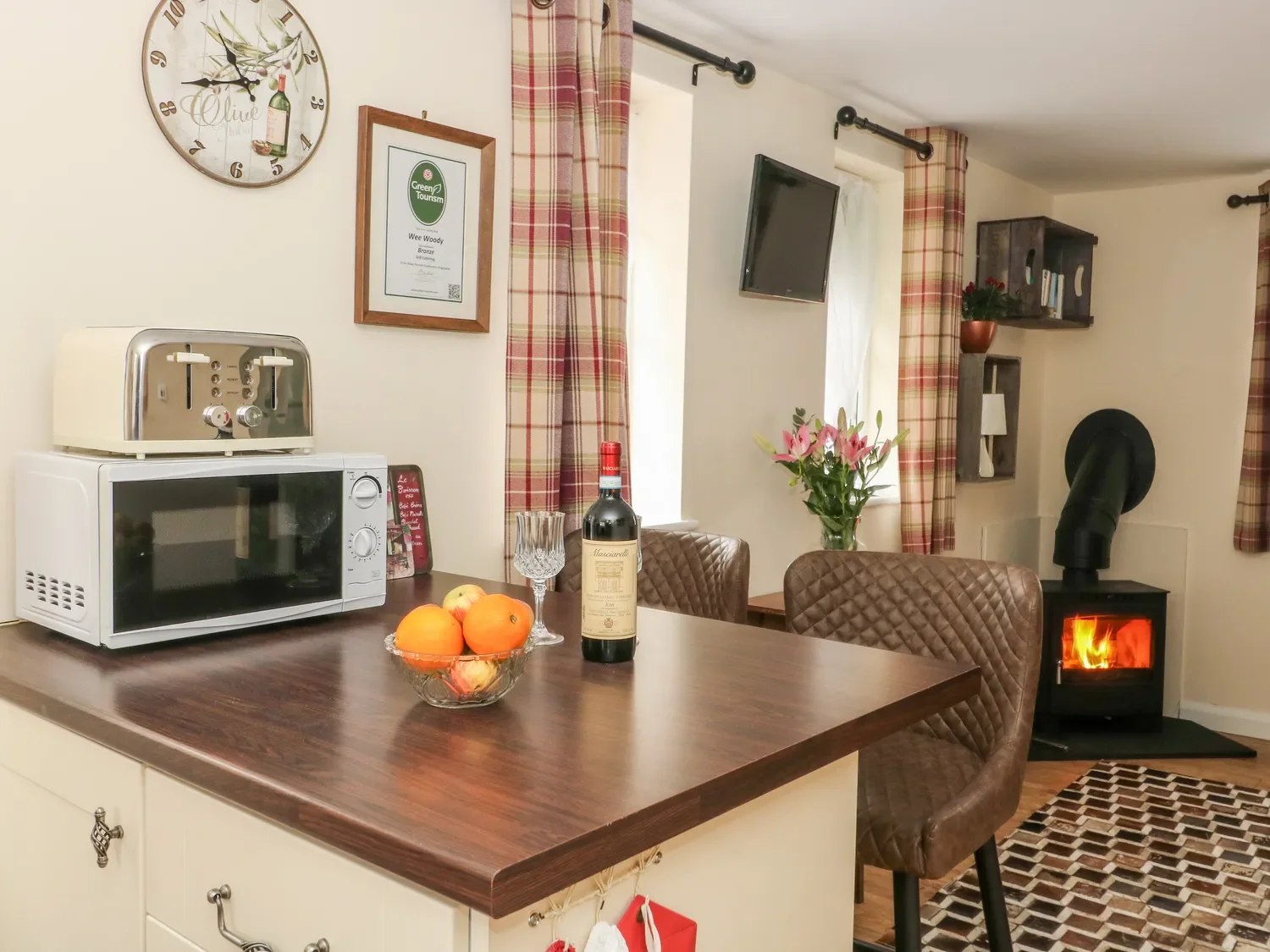 A cozy kitchen and dining area with a wooden table, fruit bowl, wine bottle, and glass. There are plaid curtains, a wall clock, a framed certificate, a microwave, a toaster, a TV, a wood stove with a visible fire, and a vase with pink lilies.