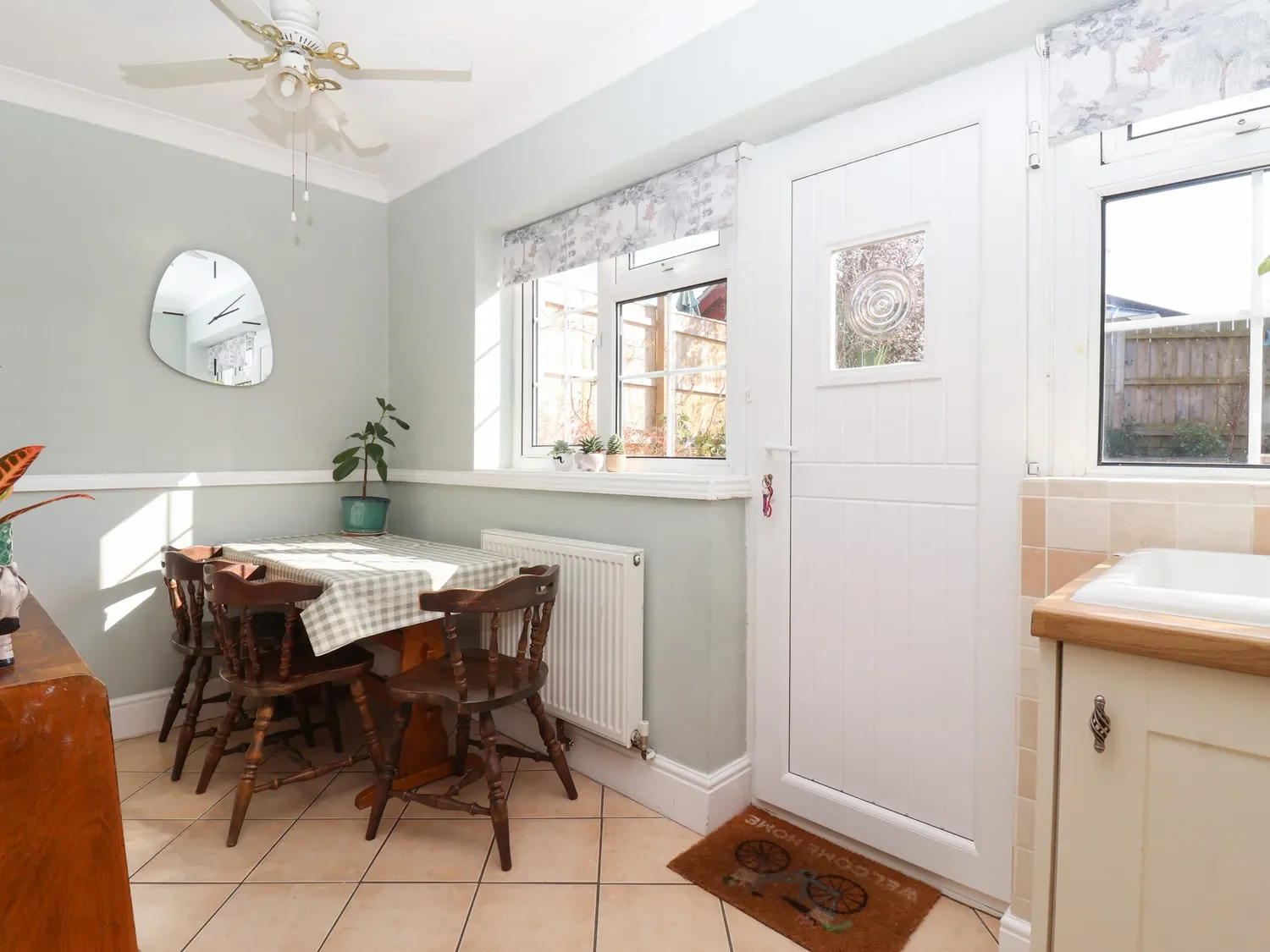 A bright kitchen corner with a small wooden table covered by a checkered cloth, surrounded by four wooden chairs, a window with a floral valance, and decorative plants on the windowsill. Sunlight casts shadows on the tiled floor.