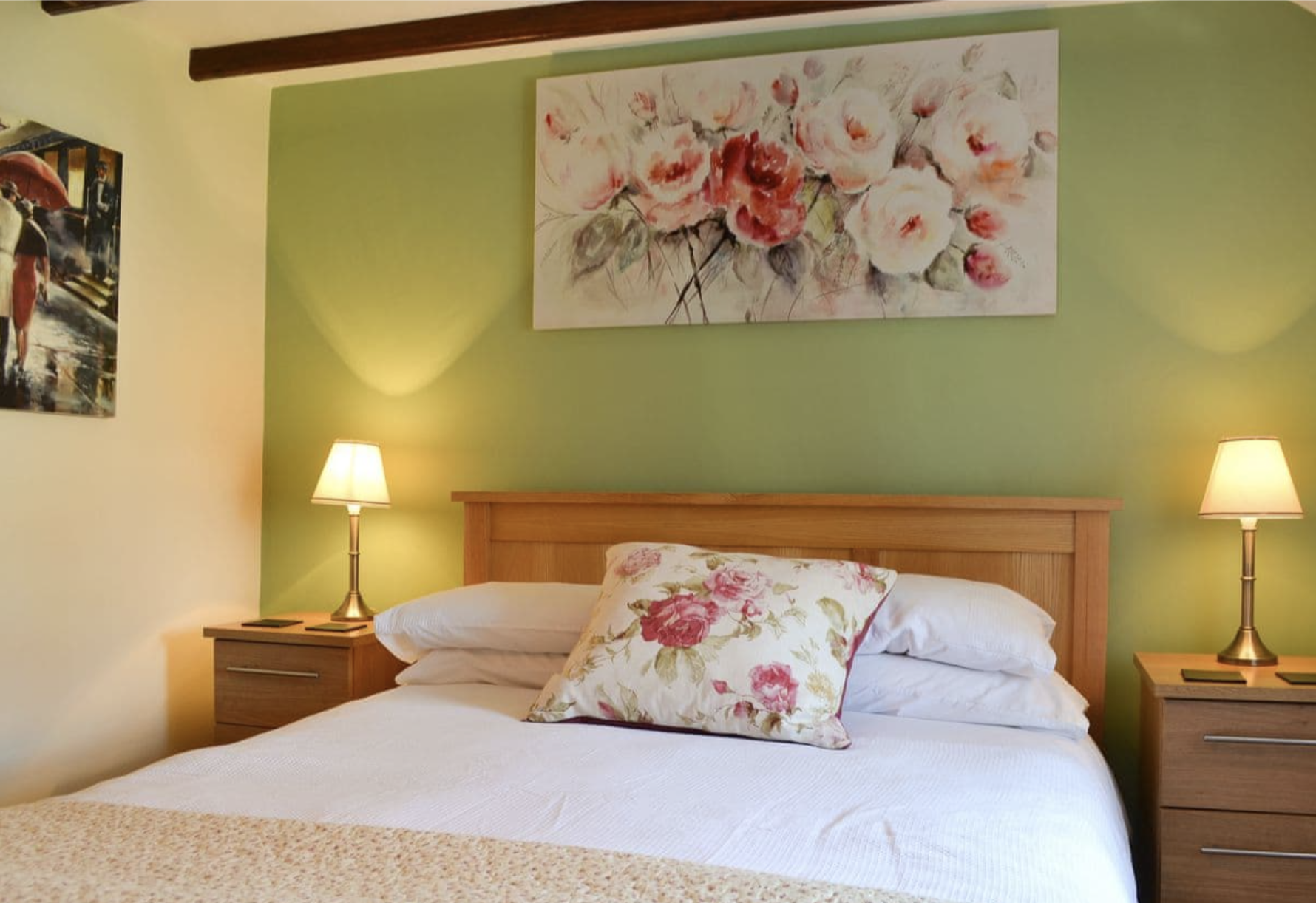 A bedroom with a wooden bed and floral bedding, green accent wall, two bedside lamps, and floral artwork above the bed.