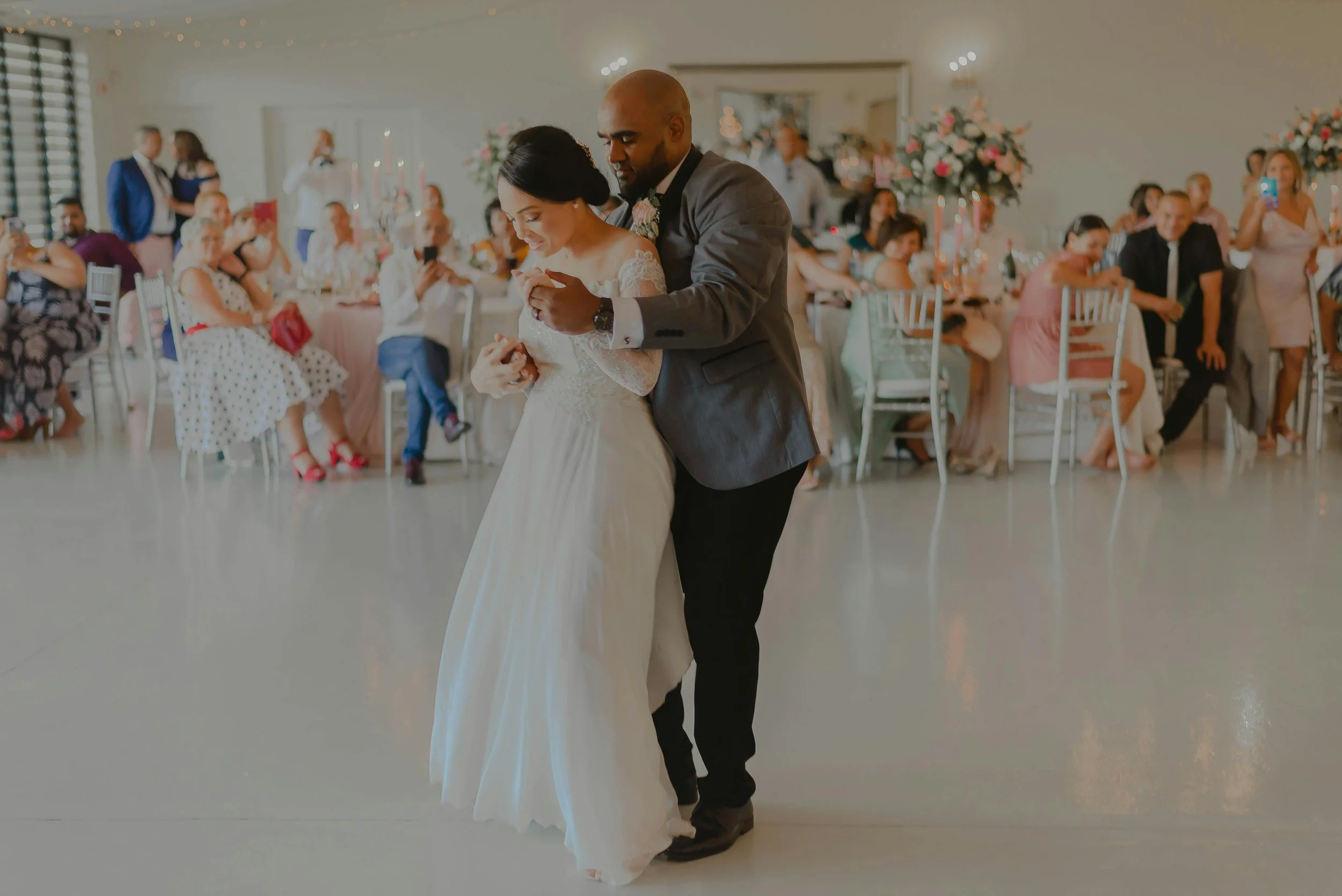 A bride and groom dance closely at their wedding reception, surrounded by seated guests taking photos and videos, with floral arrangements and soft lighting in the background.