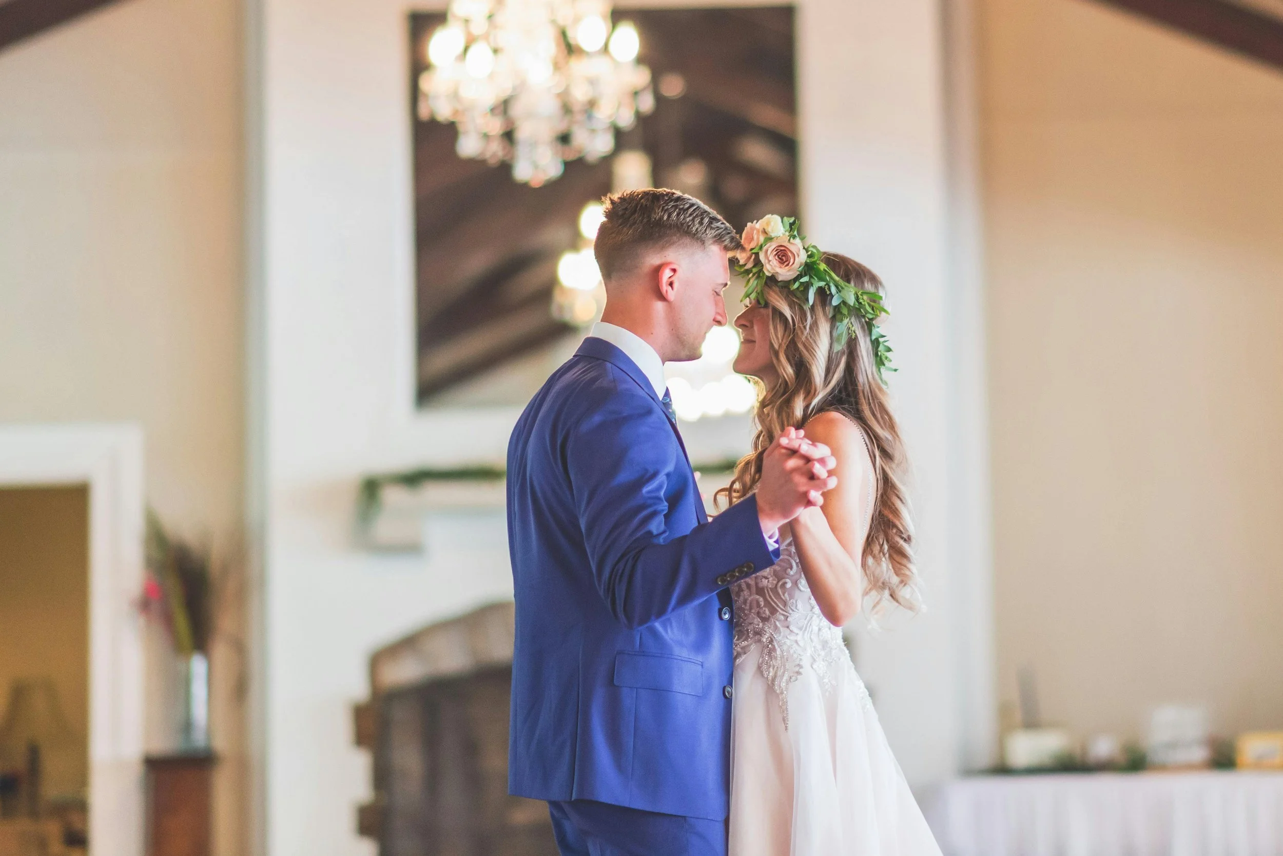 A bride and groom dance closely indoors, with the groom in a blue suit and the bride wearing a floral crown, during their wedding celebration.