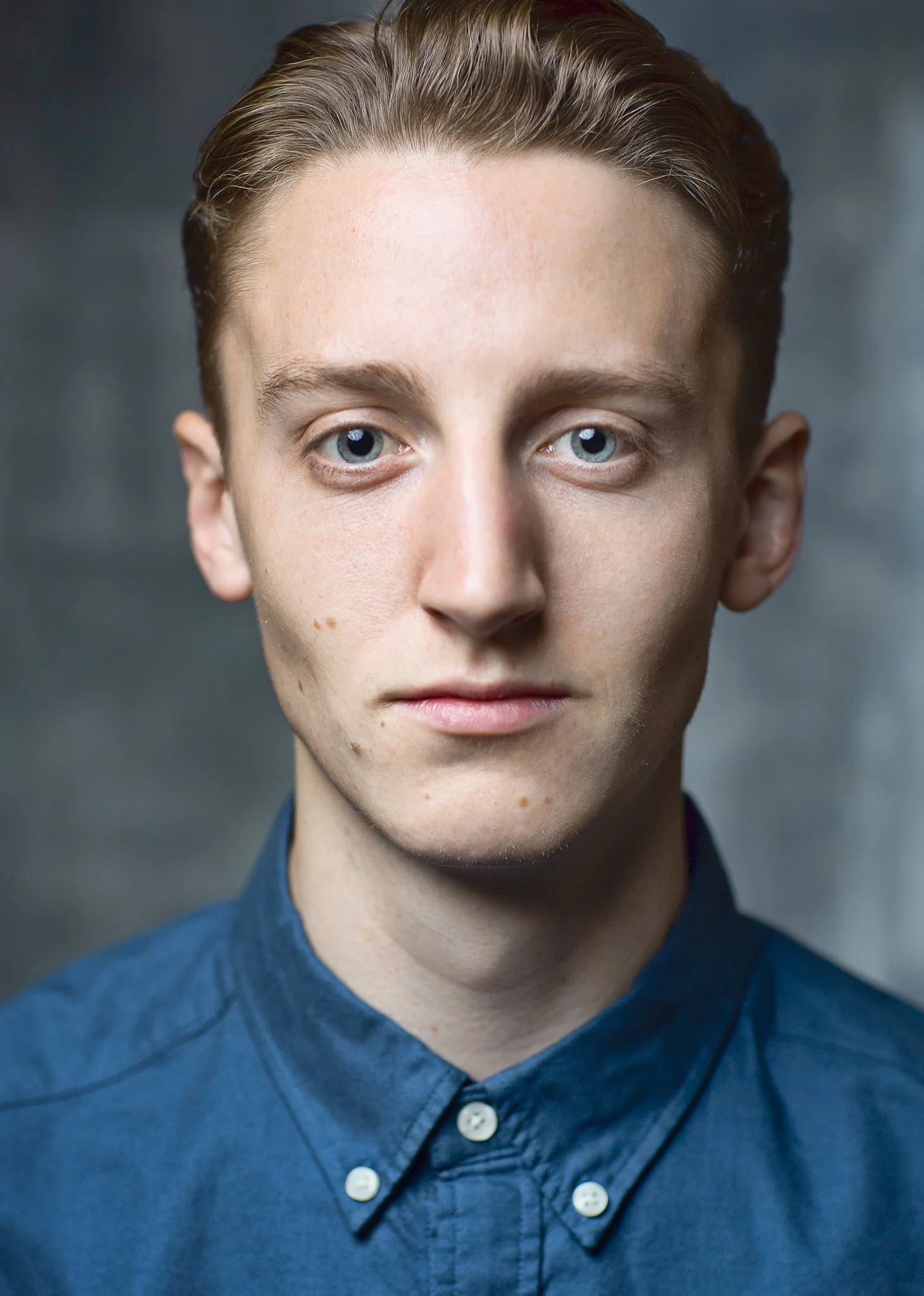 Close-up portrait of a young man with short, light brown hair, blue eyes, and fair skin, wearing a navy blue button-up shirt