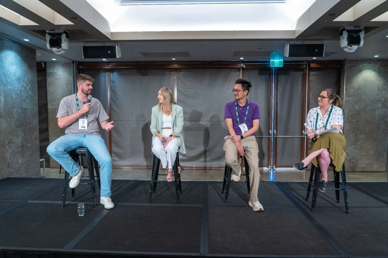 Four people sitting on bar stools on a stage, engaged in a panel discussion or interview, with a man holding a microphone speaking while the others listen.