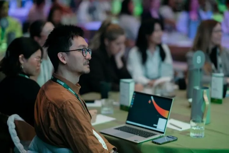 A man with glasses and a brown shirt sitting at a conference table with a laptop in front of him, surrounded by other attendees in a large conference room.